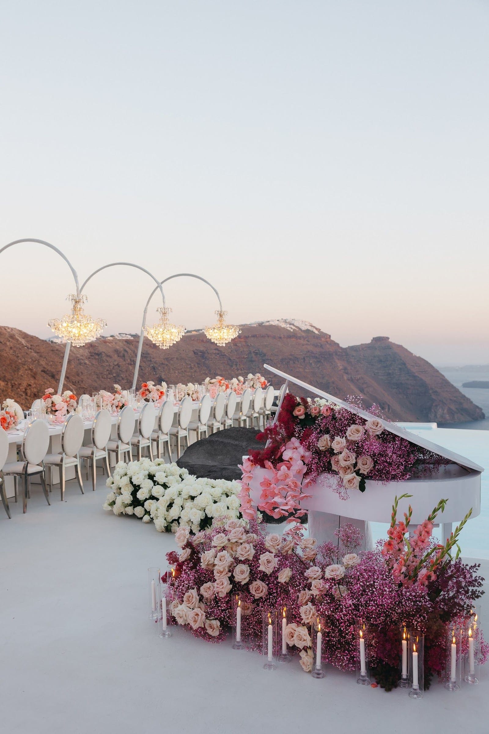 A white grand piano on a pale terrace at Aenaon Villas in Santorini, Greece, its lid and base wrapped in pink and blush roses, baby's breath and gladioli, with candles in clear glass holders along the floor. Behind it, a long reception table set with white chairs and pink floral centrepieces runs toward the edge, beneath three arched frames hung with crystal chandeliers. Soft pink dusk light falls over the caldera cliffs and sea, making this a wedding flower display of romantic, blush-toned elegance.