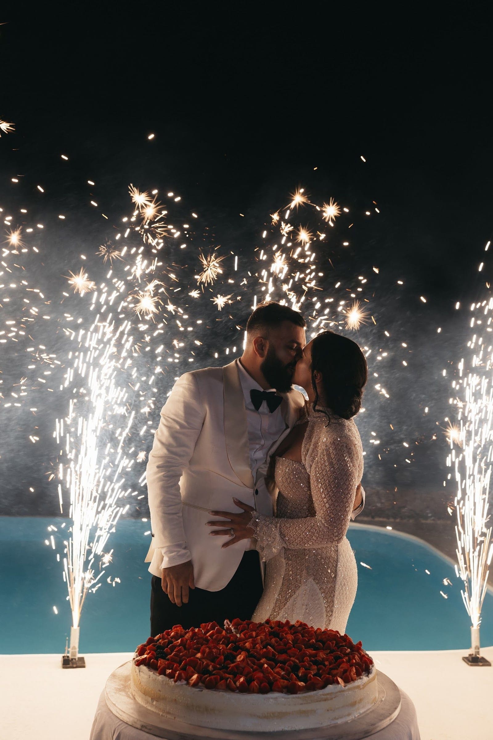 A bride and groom share a kiss at night behind a cake heaped with fresh strawberries, while tall cold-spark fountains throw glittering white sparks around them against a dark sky. He wears a white dinner jacket with a black bow tie, and she wears a sparkling, long-sleeved gown. Beyond them a turquoise pool glows at Aenaon Villas in Santorini, Greece, in this wedding celebration.
