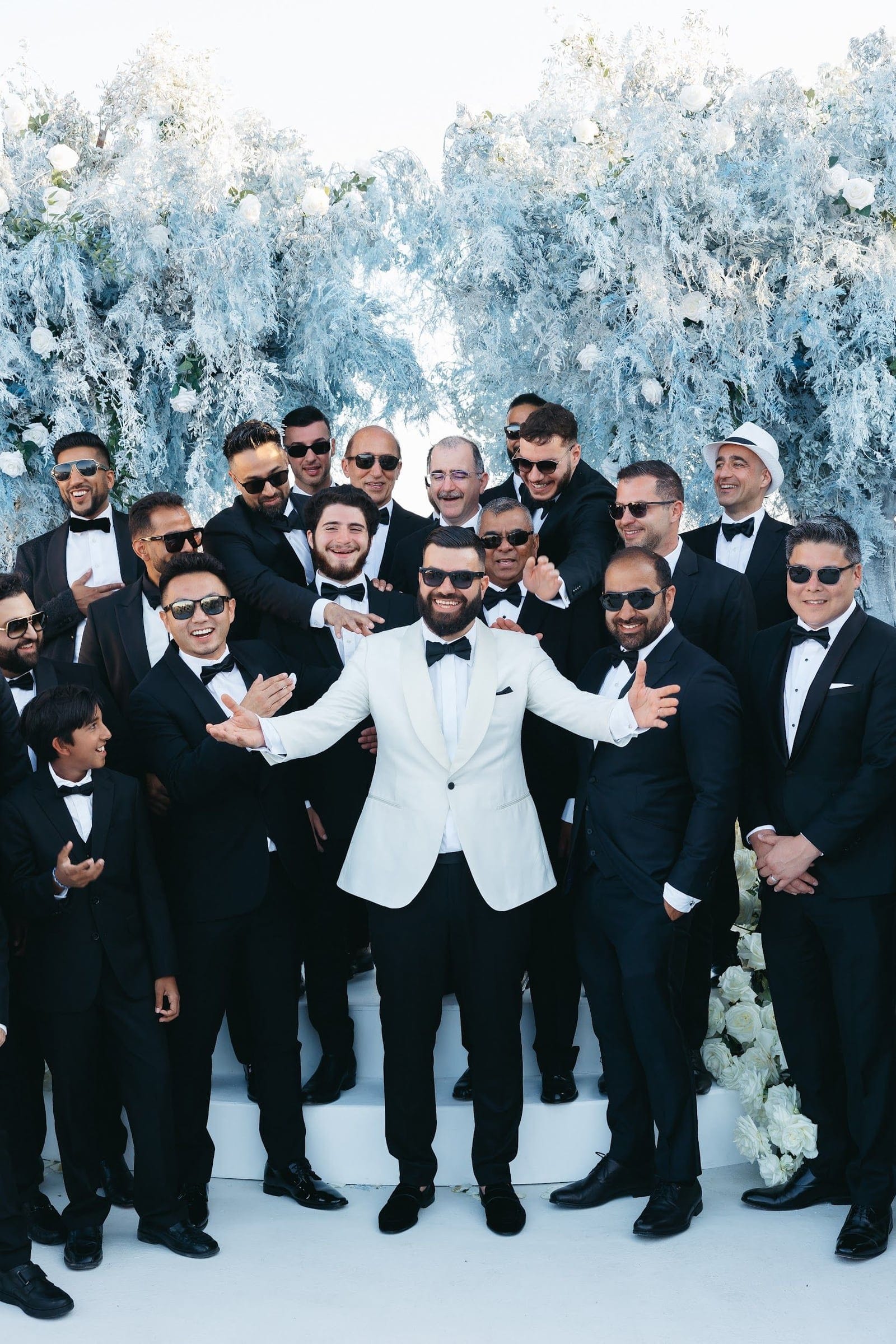 A bearded groom in a white dinner jacket and black bow tie stands at the centre of a crowd of smiling men in black tuxedos and sunglasses, arms spread wide in welcome. Behind them a tall arch is wreathed in pale blue-grey foliage, likely dusty fern-like branches, dotted with white roses, forming a striking wedding backdrop at Aenaon Villas in Santorini, Greece.