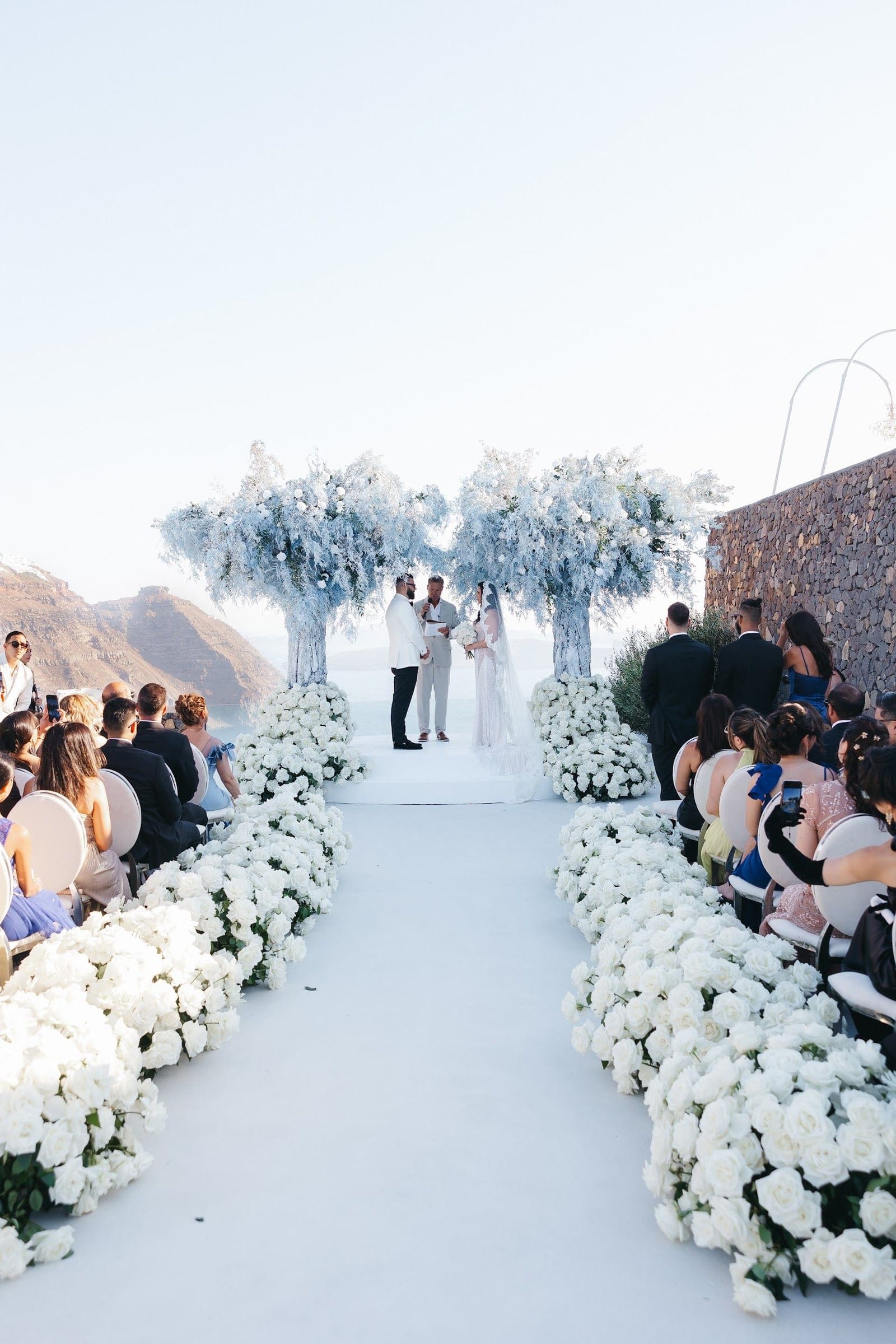 A wedding ceremony at Aenaon Villas in Santorini, Greece, framed by two tall, airy arches of pale blue-grey foliage with white blooms. A white aisle is lined on both sides with dense rows of white garden roses leading to the couple, who stand beneath the arches facing an officiant, the bride in a long veil and the groom in a white jacket. Guests in dark suits and soft pastels sit on white chairs along the path, all beneath a hazy, pale sky over the caldera.