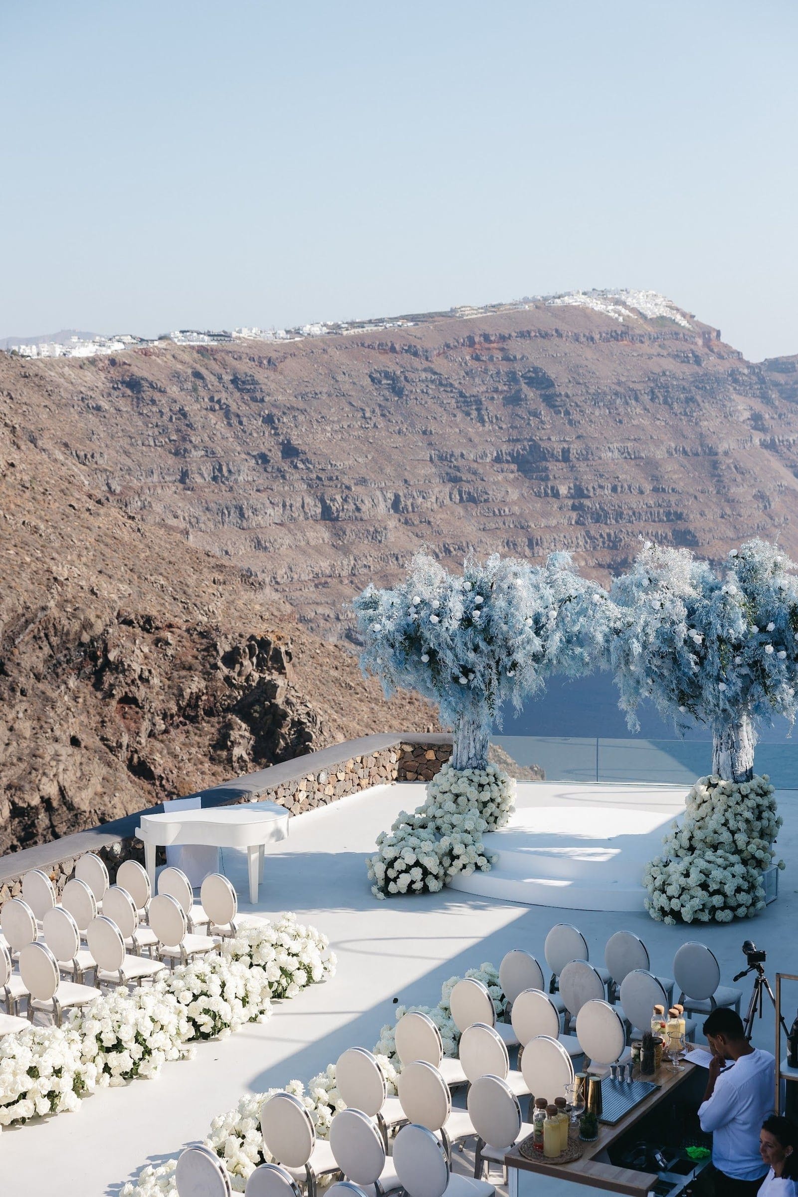 A wedding ceremony set on a white terrace at Aenaon Villas in Santorini, Greece, with two tall arches draped in pale blue and white flowers and foliage, their bases banked with white roses. White chairs with round backs line both sides of an aisle edged with white blooms, a white grand piano sits to the left, and the caldera cliffs and sea stretch out behind under a clear, hazy sky.