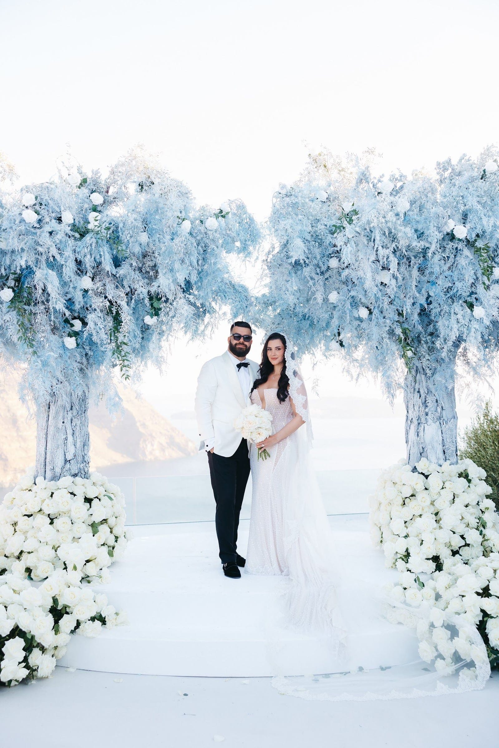 A bride and groom stand side by side under an arch of pale blue, feathery foliage dotted with white roses, with a heap of white roses spilling across the floor at each side, at a wedding at Aenaon Villas in Greece. He wears a white dinner jacket with a black bow tie and dark sunglasses, and she wears a fitted, sparkling gown with a lace-edged veil that trails behind her, holding a white rose bouquet. Hazy sea and cliffs fill the background, giving the scene a soft, airy calm.