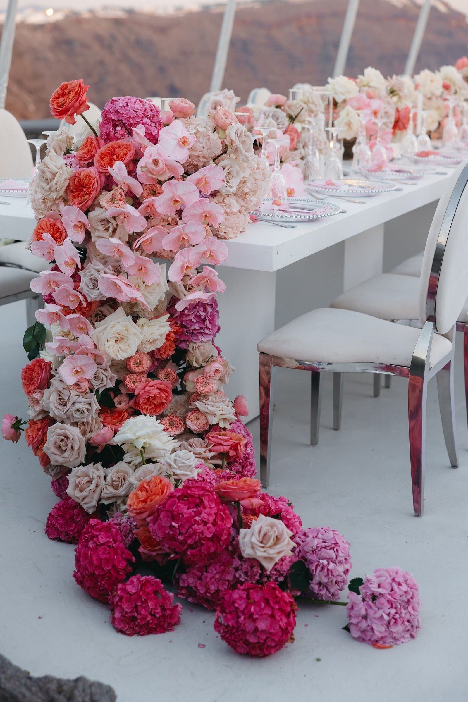 A long white dining table on a clifftop terrace at Aenaon Villas in Santorini, Greece, is dressed with a cascading wedding table installation in pink and coral. Ombré garden roses in peach, cream and coral, pink phalaenopsis orchids, and magenta and pale pink hydrangeas tumble down the side of the table and pool across the pale floor. Pale plates and glassware line the table beyond, with mirrored chairs in the foreground and the caldera and sea softly visible behind.