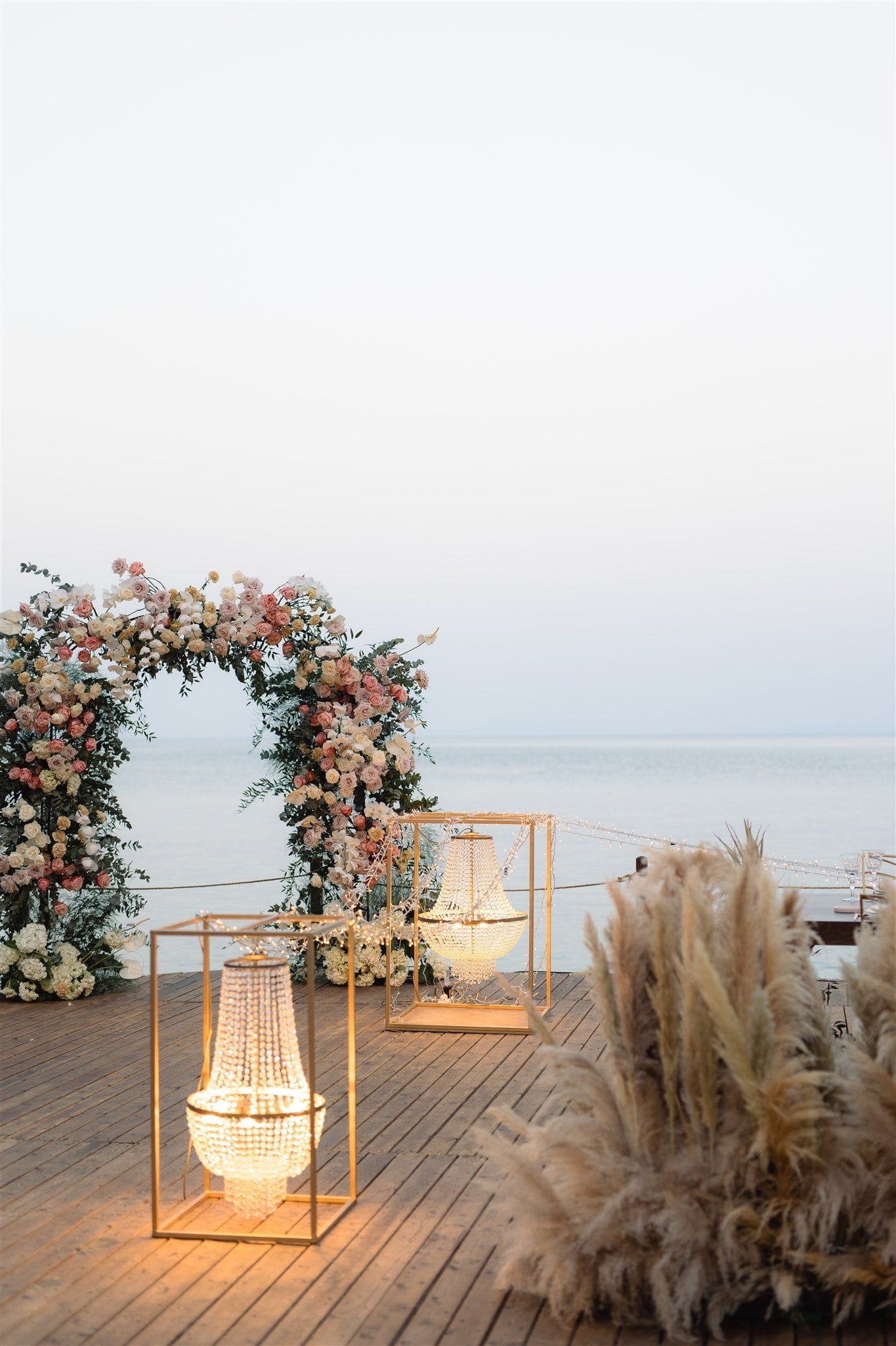 A round wedding arch of blush and cream roses, white hydrangeas and sprigs of eucalyptus stands on a wooden deck at Lazarou Beach in Greece, with the calm sea stretching out behind it under a pale evening sky. Two gold cube frames holding glowing crystal chandeliers flank the path, and a clump of soft pampas grass sits to the right. The warm light against the cool, hazy blue creates a serene, romantic mood for a seaside ceremony.