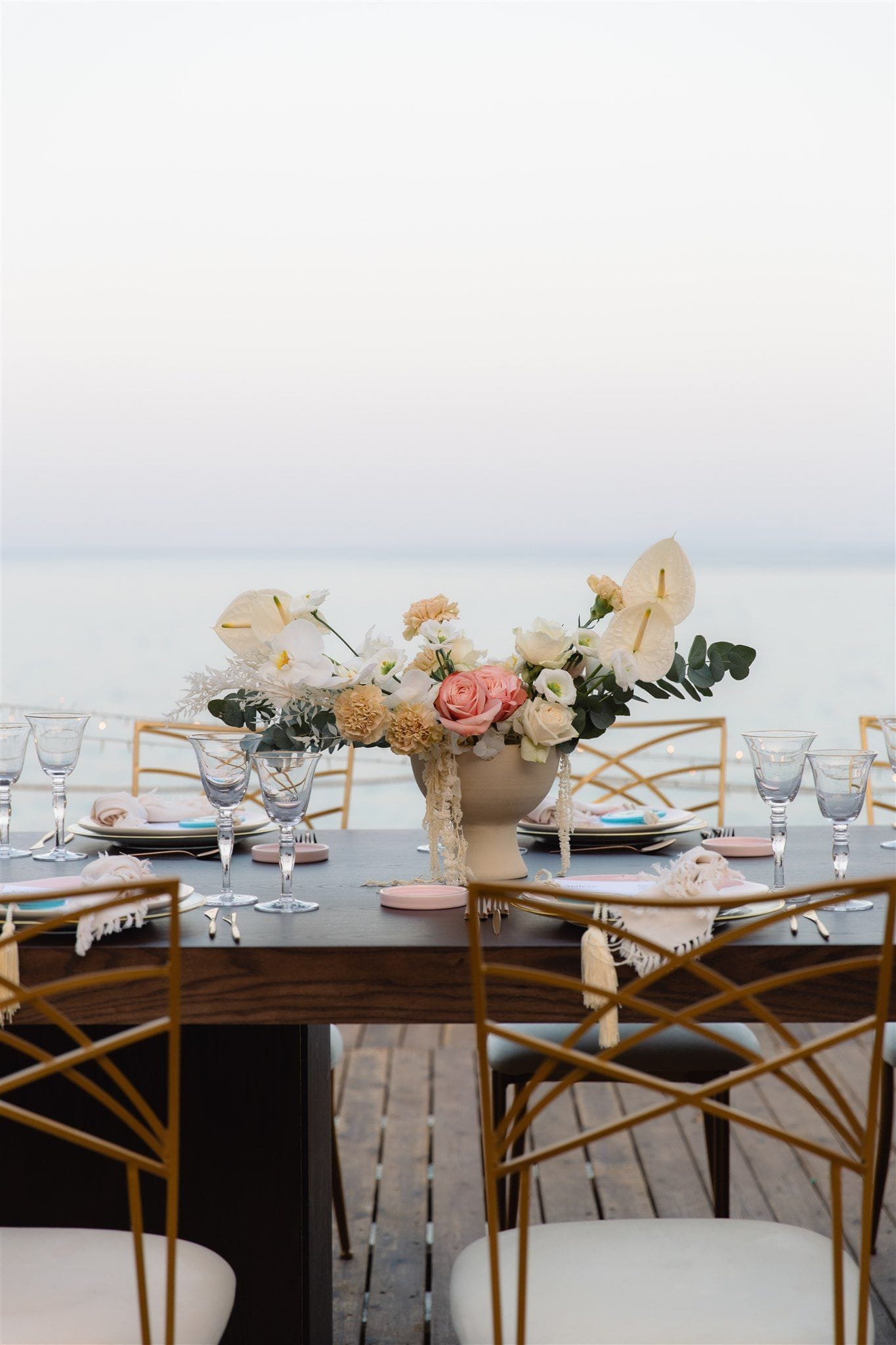 A long dark wooden table set for a wedding dinner on a wooden deck at Lazarou Beach in Greece, with the calm sea and pale sky stretching behind it. At the centre sits a low cream vase holding white anthurium, peach carnations, pale roses, and sprigs of eucalyptus, with trailing amaranthus hanging down its side. Clear stemmed glasses, cream napkins with tassels, and blush-pink coasters line the table, and gold geometric chairs frame the soft, misty evening scene.