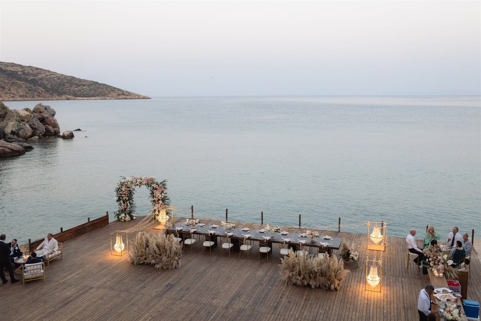 A long wooden deck at Lazarou Beach in Greece overlooks a calm, pale sea, with a rocky headland on the left. A long dark wedding table is set for a dinner, its place settings lit by lanterns and strings of fairy lights, with a pink and cream floral arch of roses and foliage at the far end and tall dried pampas grass clusters flanking the table. Guests sit in soft lamplight at dusk.