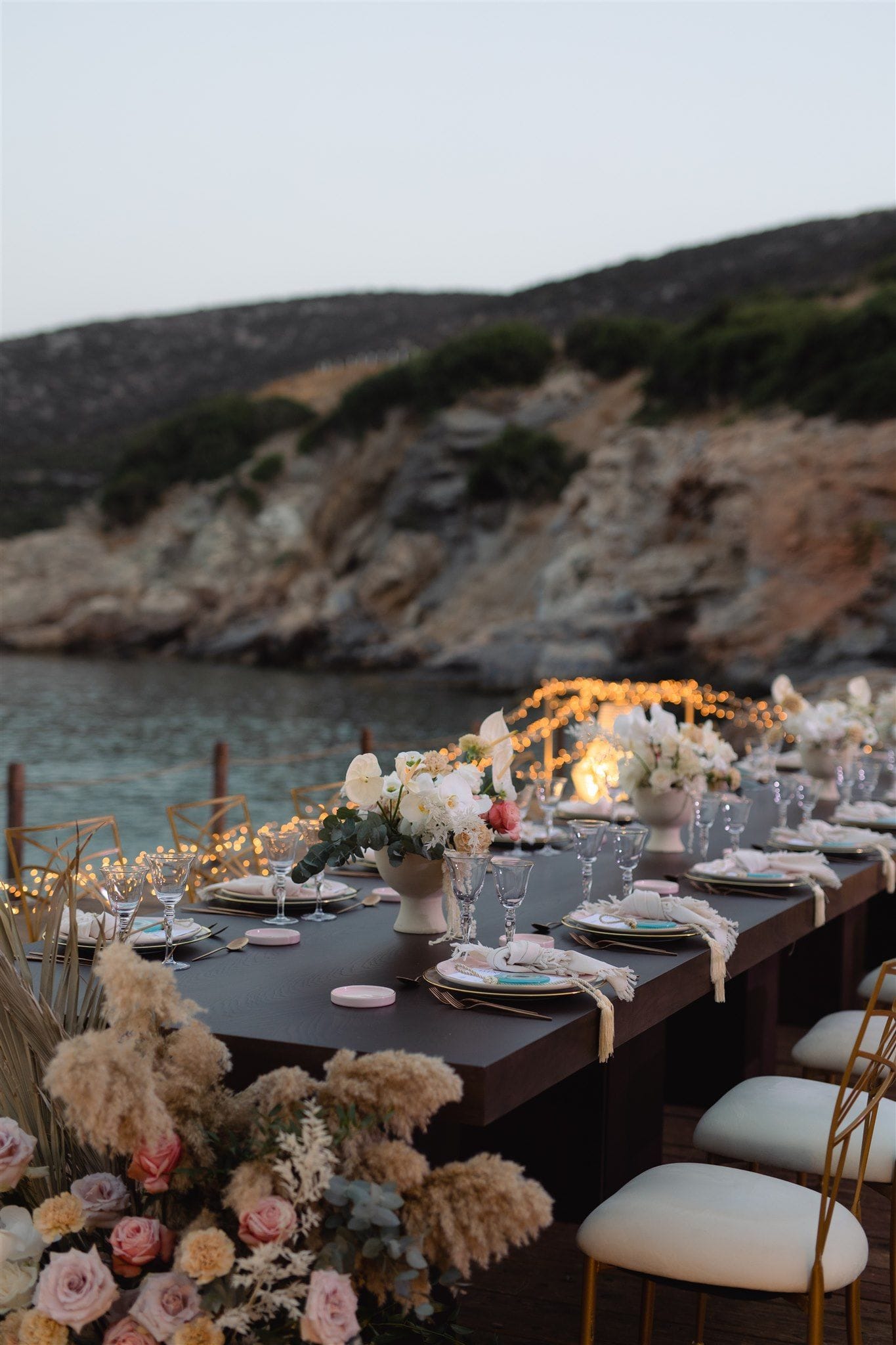 A long dark wooden wedding table set for dinner at dusk on a seaside terrace at Lazarou Beach in Greece, with white plates, gold cutlery, and white napkins tied with fringed ends. Centrepieces of white anthuriums, roses and eucalyptus rise from pale vases, while low arrangements of blush roses, dried pampas grass and soft ivory blooms fill the foreground. Fairy lights glow warmly along the edge of the calm water, and rocky, scrubby hills rise behind in the soft evening light.