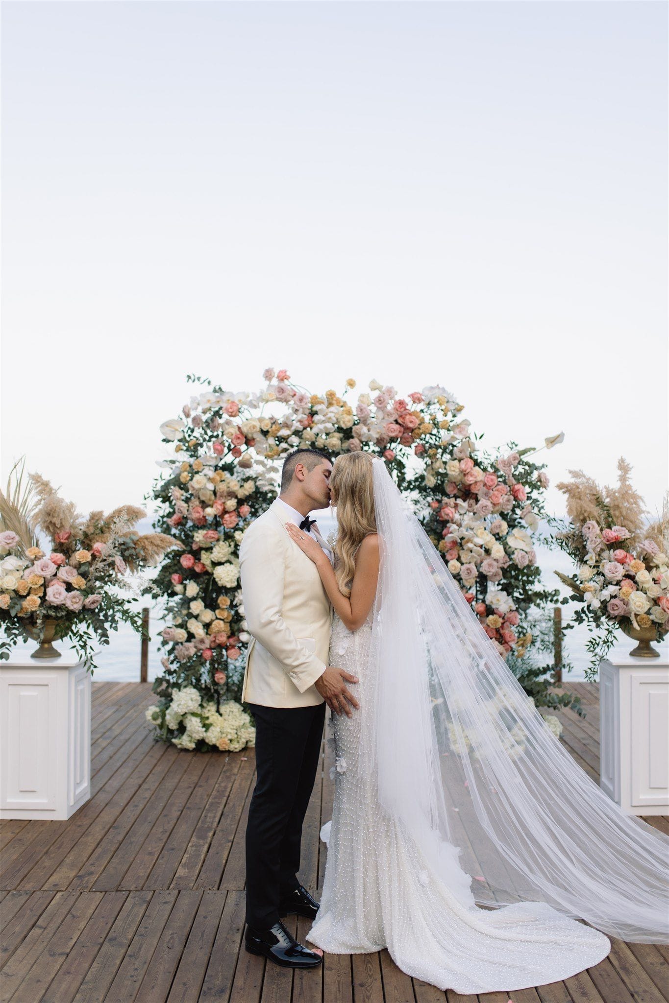A couple shares a tender kiss in front of a full floral arch of pink, cream and peach roses, white hydrangeas and foliage, with pampas grass urns flanking them on a wooden deck by the sea. The groom wears a cream dinner jacket with a black bow tie, and the bride wears a pearl-beaded gown with a long, flowing veil. This is wedding flowers at Lazarou Beach in Greece.