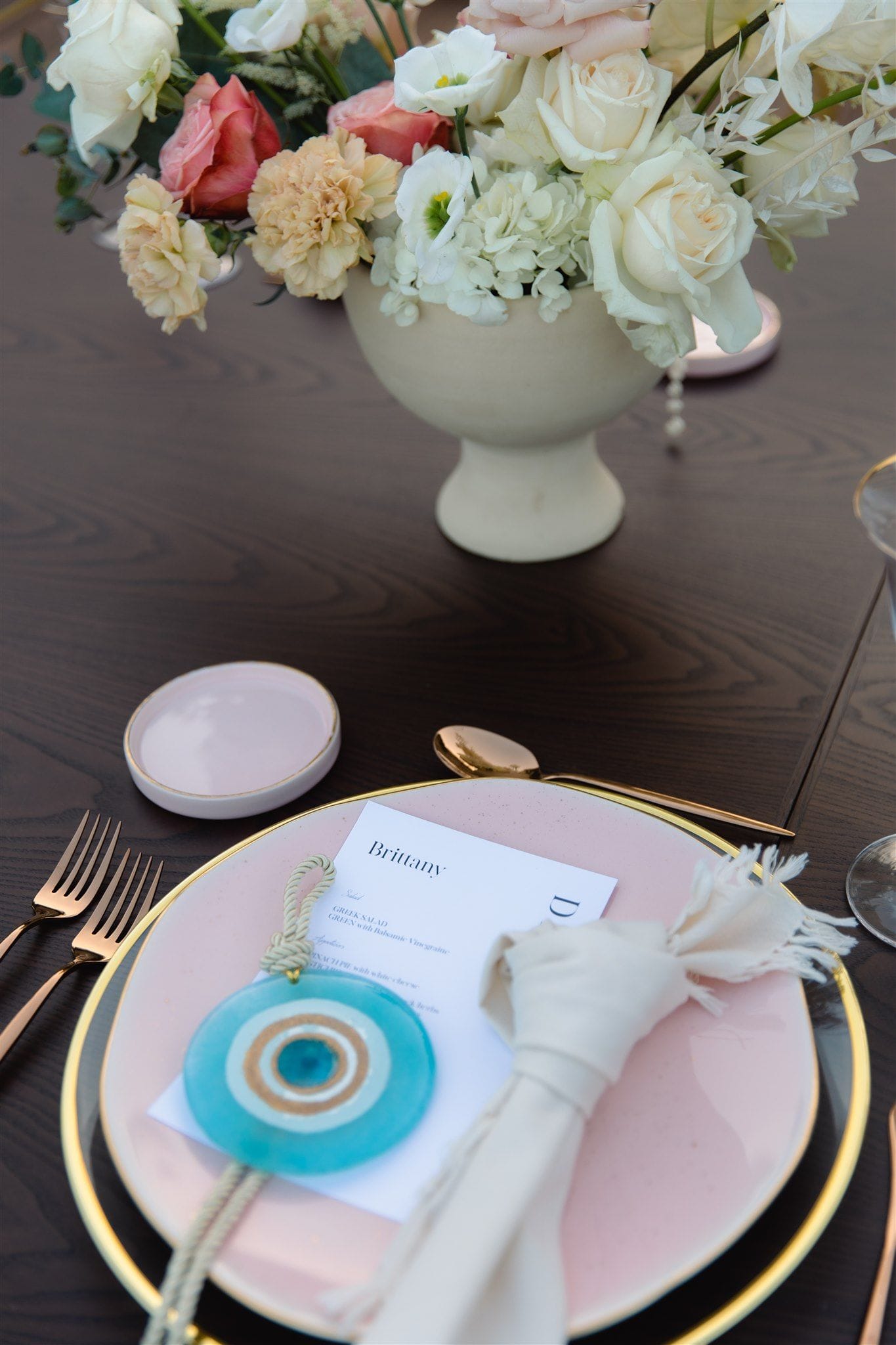 A place setting on a dark wooden table at Lazarou Beach in Greece, with a blush-pink plate rimmed in gold and a cream napkin tied with a fringed cord. On the plate sits a printed menu card reading "Brittany" and a turquoise glass evil eye charm. Behind it, a footed cream urn of white roses, ranunculus and hydrangea, with coral roses and peach carnations, forms a wedding table centrepiece.