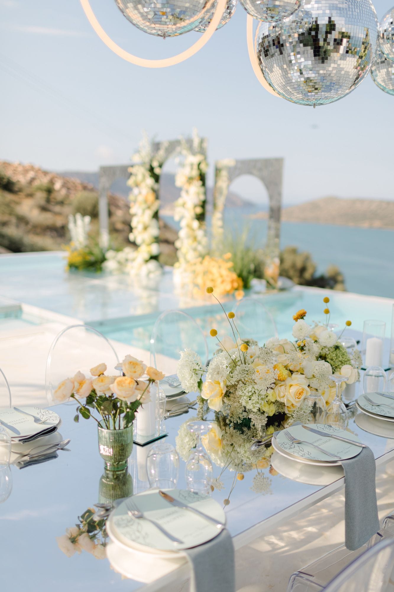 A long glass wedding table on a clifftop terrace at Dream Art Villa in Schisma Elountas, Crete, Greece, set with pale cream roses and white hydrangea-like blooms, with yellow billy buttons scattered through the centrepiece. Small vases of cream roses sit along the table beside clear acrylic chairs, grey linen napkins, and mirror-like reflections. Above hang large silver mirror disco balls, and beyond the infinity pool an arch of white and yellow blooms frames the blue Cretan sea and hills.