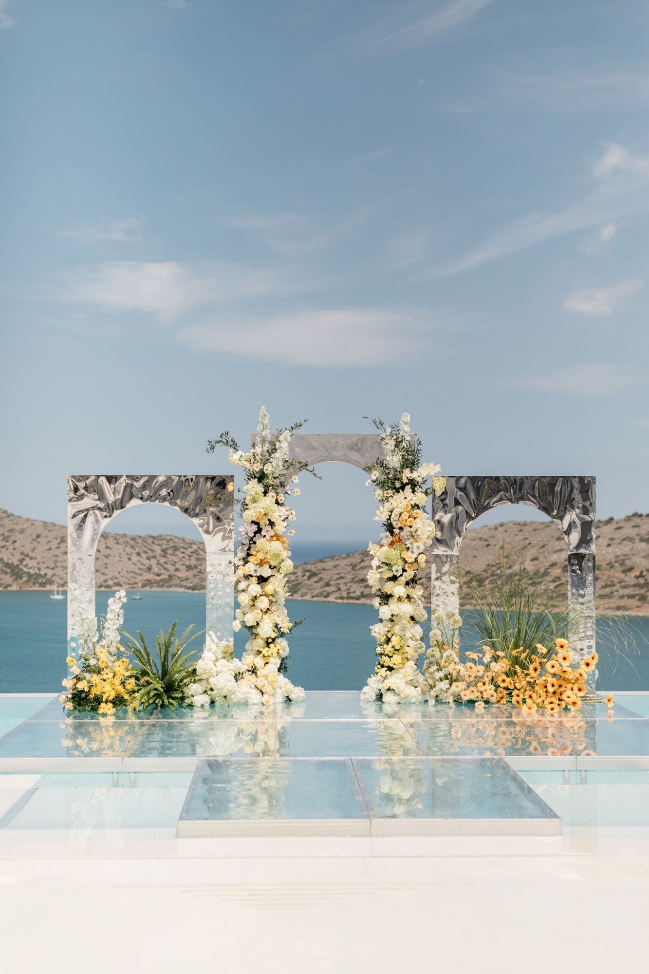 Three arched mirror-finished panels stand on a glossy glass platform above the sea, framing a wedding ceremony arch at Dream Art Villa in Schisma Elountas, Crete, Greece. Two tall columns of white and cream blooms, likely roses and hydrangea with sprigs of greenery, climb the centre arches, while clusters of yellow and apricot flowers and spiky grasses sit at their feet. Calm turquoise water and dry, rocky hills stretch behind under a pale blue sky, and the reflections in the glass floor add to the airy, elegant mood.
