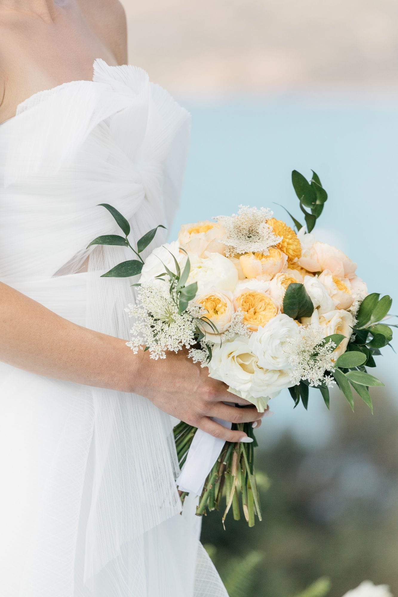 A bride in a white pleated tulle gown holds a round bouquet of apricot and cream garden roses, white ranunculus and lacy white Queen Anne's lace, with sprigs of green eucalyptus and trailing foliage, bound with a white ribbon. Soft blue sea and a blurred hillside fill the background. These wedding bouquet flowers were arranged at Dream Art Villa in Schisma Elountas, Crete, Greece.