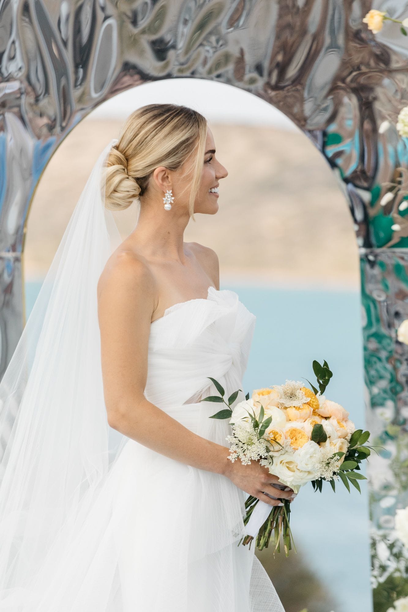 A bride in a strapless white tulle gown stands in profile, smiling toward the left, her blonde hair swept into a low chignon with a sparkling drop earring. She holds a bridal bouquet of pale yellow garden roses, white roses and cream lace-like flowers, accented with green eucalyptus sprigs. Behind her is a mirrored, rippled arch with pale blue sea and soft golden hills beyond, at Dream Art Villa in Schisma Elountas, Crete, Greece.
