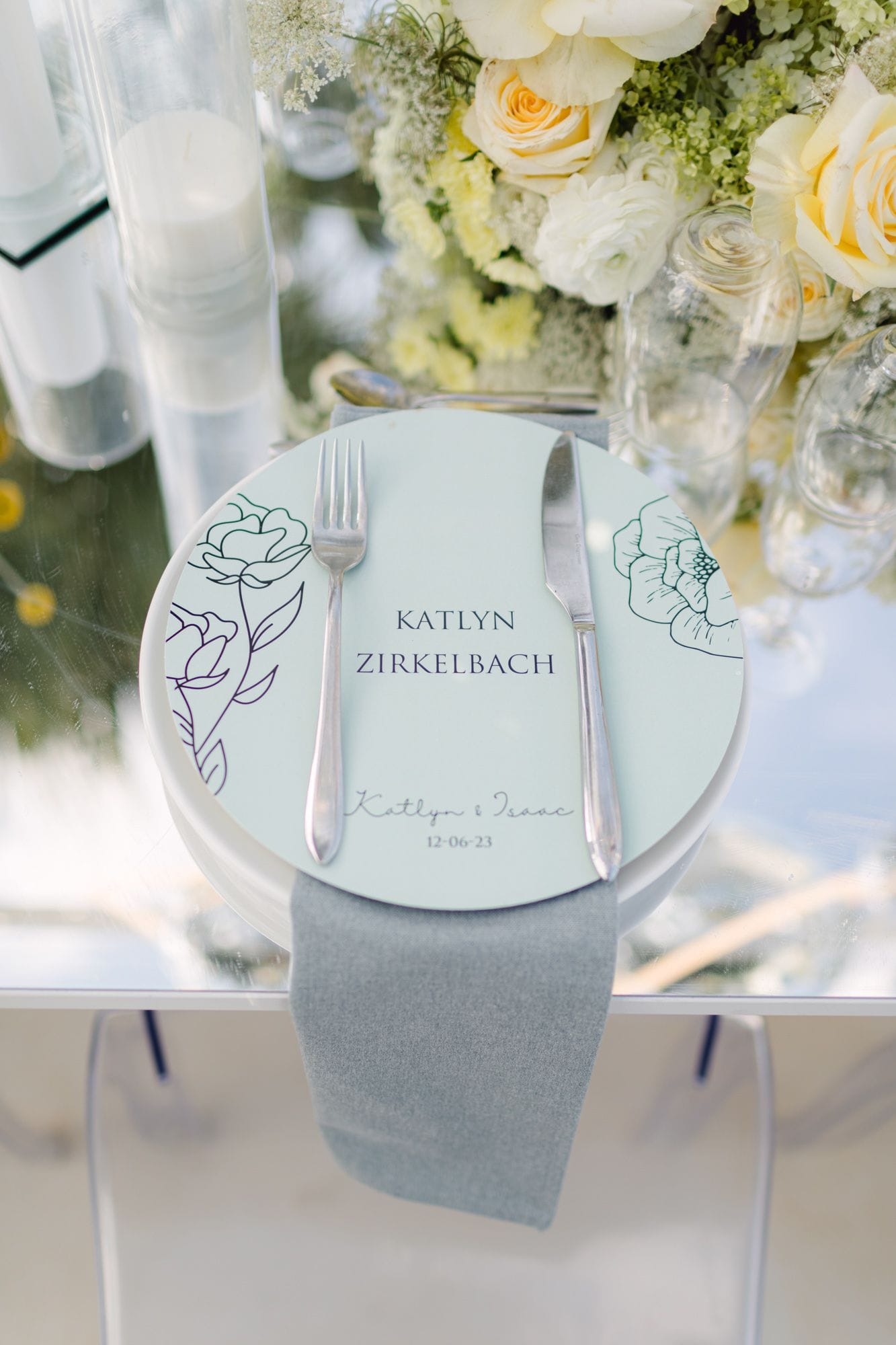 A round pale-mint place card with fine line drawings of peony-like flowers in deep teal, framing the names Katlyn Zirkelbach and "Katlyn & Isaac", dated 12-06-23, set on a white plate beside a silver fork and knife and a grey linen napkin on a glass table. Behind it, pale yellow garden roses, white ranunculus and cream blossom in a lush centrepiece, at a wedding table in Schisma Elountas, Crete, Greece.