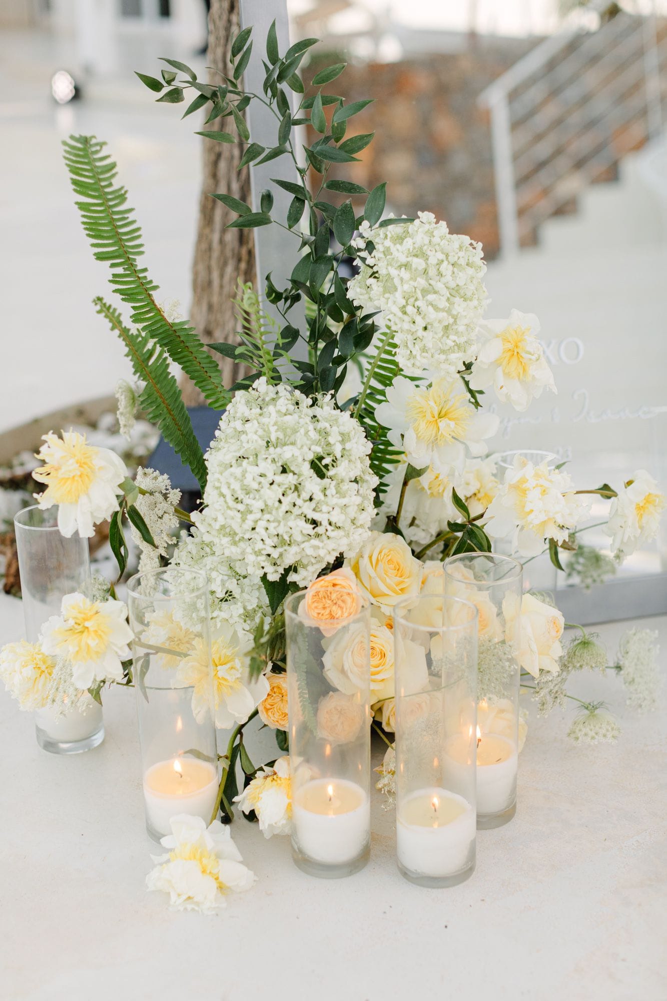 A low wedding arrangement on a pale stone terrace at Dream Art Villa in Schisma Elountas, Crete, Greece, built from white hydrangea heads, cream and butter-yellow peonies, and soft apricot garden roses. Tall green fern fronds and sprigs of eucalyptus rise behind them, and clear glass cylinders holding lit white candles stand at the base, their warm glow catching the petals.