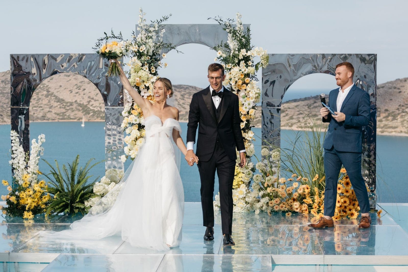 A bride in a white off-the-shoulder gown raises a yellow and cream bouquet in triumph as she walks hand in hand with her groom in a black tuxedo along a glossy platform at Dream Art Villa in Schisma Elountas, Crete, Greece. Behind them, two polished silver arches frame an altar of white and yellow flowers and greenery, with a calm blue bay and dry hills beyond. The wedding flowers are arranged in tall sweeps with yellow daisies at the base, and a celebrant in a navy suit smiles to the side.