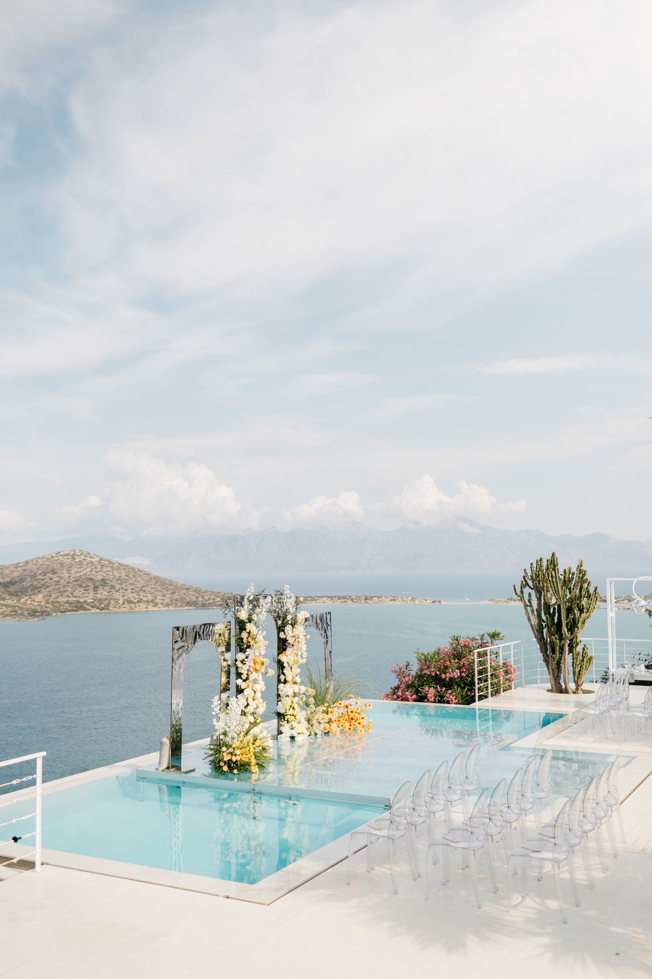 A wedding ceremony set at the edge of an infinity pool at Dream Art Villa in Schisma Elountas, Crete, Greece, with a pair of dark mirrored arches draped in white orchids and garden blooms and yellow flowers at their base. Rows of clear acrylic Ghost chairs face the arch across the pale turquoise water, with the blue Aegean, a rocky headland and distant mountains stretching behind under a soft, hazy sky.