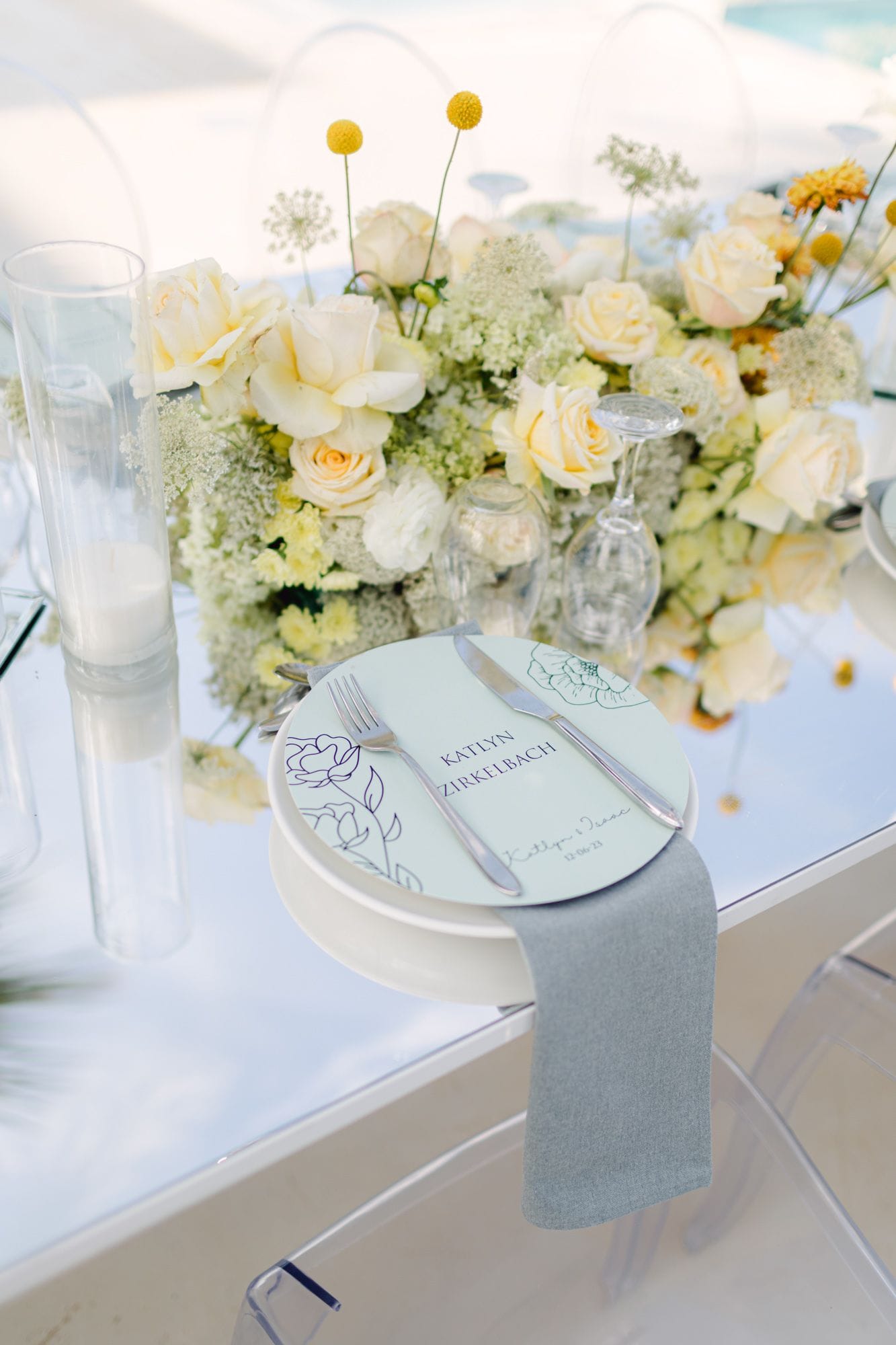 A wedding table setting at Dream Art Villa in Schisma Elountas, Crete, Greece, with a pale cream and yellow centrepiece of garden roses, white hydrangea-like blooms, Queen Anne's lace and yellow craspedia balls rising above a clear glass table. In front, a pale blue charger carries a name card with delicate navy line-drawn peonies, set beside silver cutlery and a grey-blue linen napkin.