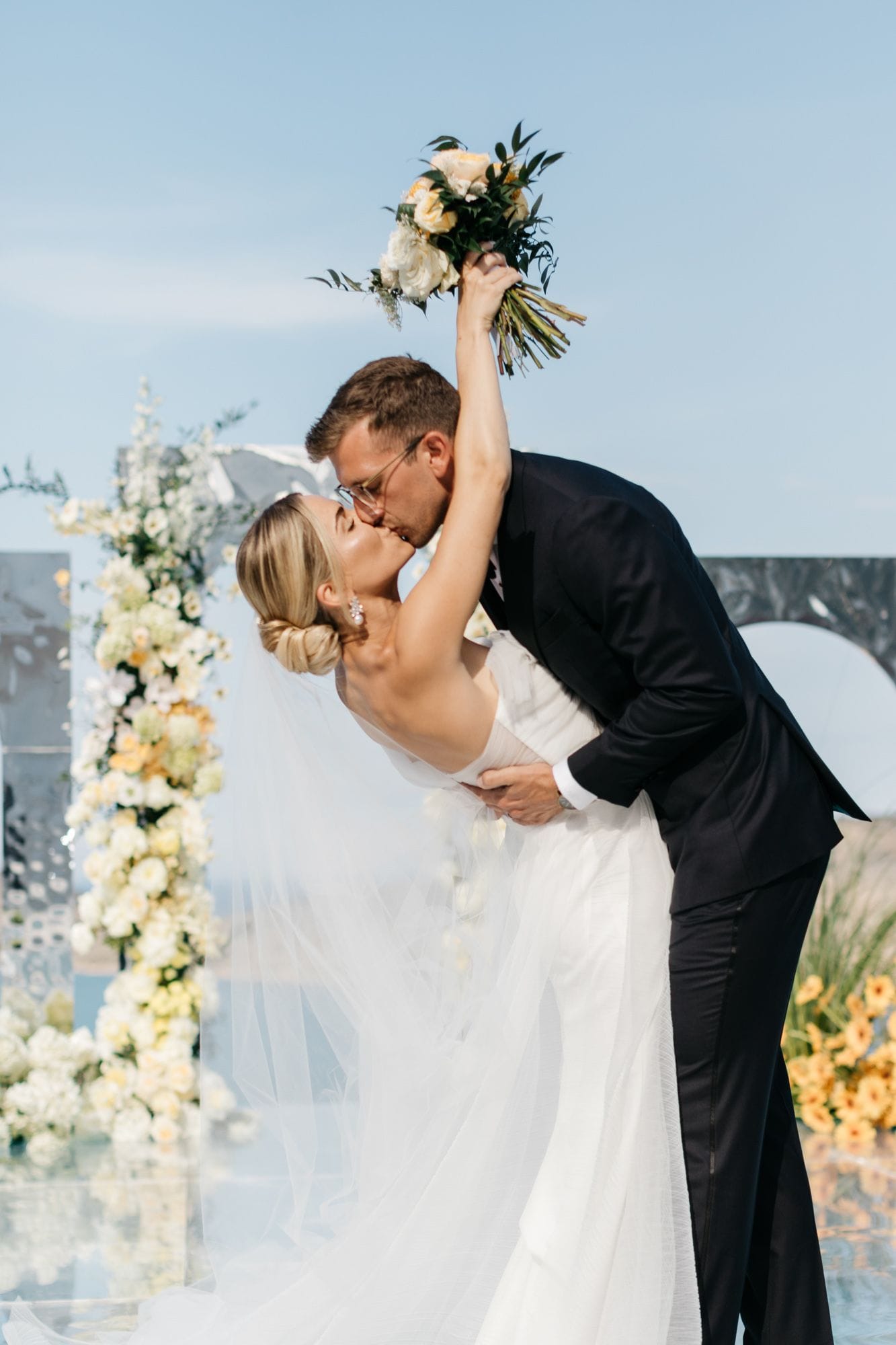 A couple shares a dramatic dip-and-kiss beneath a clear blue sky, the bride's arm raised high and still holding her bouquet of greenery and pale stems. Behind them stands a wedding arch wrapped in a cascading column of white and cream blooms, with soft yellow flowers and hydrangea-like clusters, all set against a light stone backdrop at Dream Art Villa in Greece. The bride wears a white tulle gown and a sheer veil that flows across the frame, and the groom wears a dark suit and clear-framed glasses.