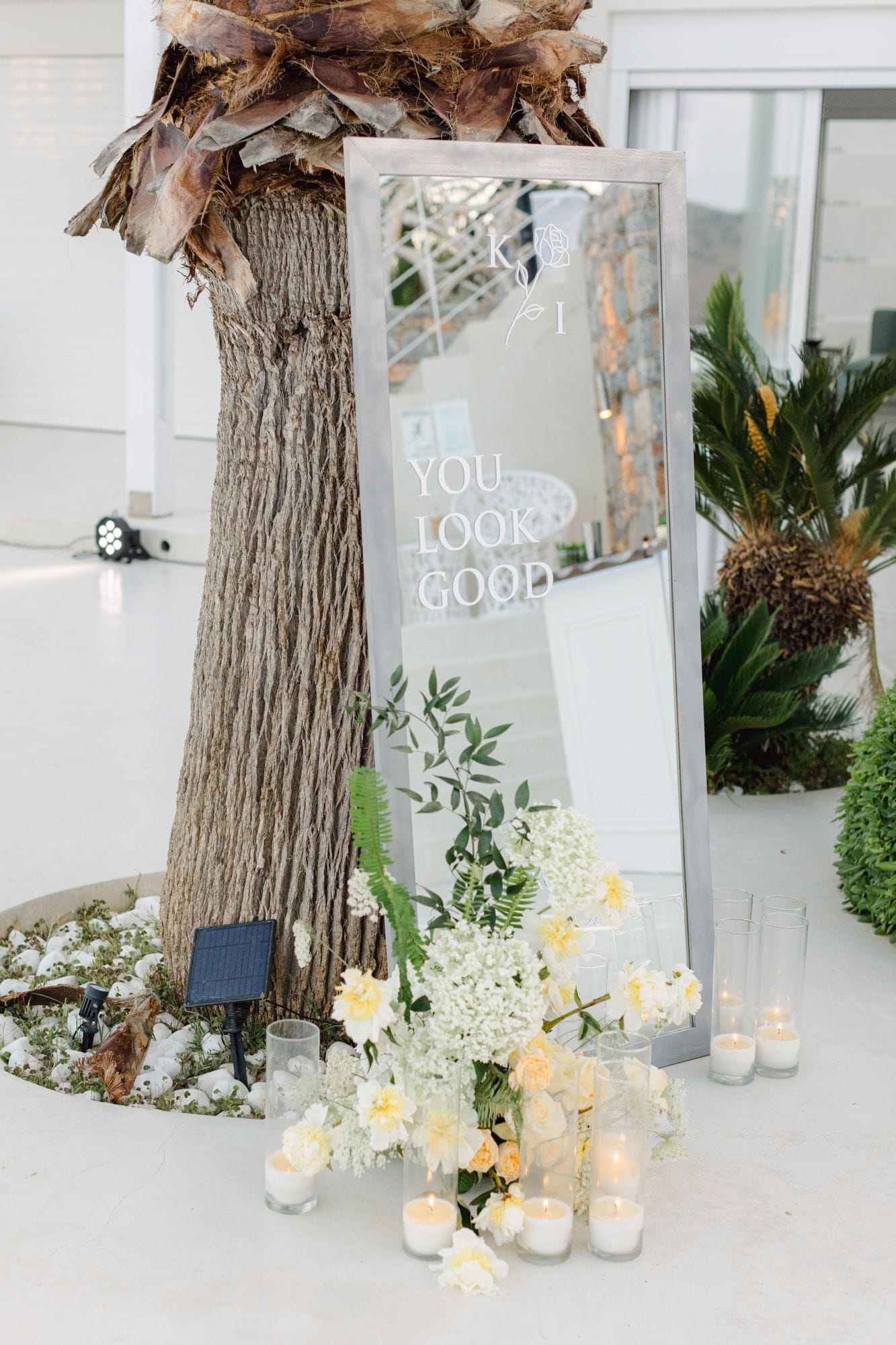 A tall silver-framed mirror leans against the trunk of a palm tree on a white terrace at Dream Art Villa in Schisma Elountas, Crete, Greece, with "YOU LOOK GOOD" etched across the glass and a small rose monogram "K & J" above it. At the mirror's base sits a wedding arrangement of cream and pale yellow roses, white hydrangea, gypsophila, and green fern and eucalyptus sprigs, flanked by clear glass cylinders holding lit white candles. A solar spike and a scatter of white stones surround the palm's base, and the soft daylight gives the whole scene a calm, airy feel.