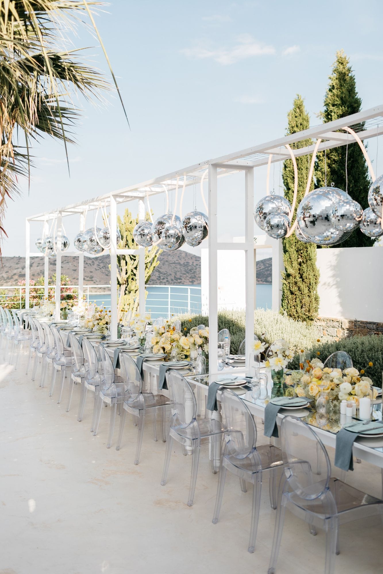 Long wedding tables on a sunlit terrace at Dream Art Villa in Schisma Elountas, Crete, Greece, set with clear acrylic chairs, dusty blue linens and centrepieces of pale yellow and cream roses and ranunculus. Silver mirror balls hang in clusters from a white pergola above, and a palm frond and cypress trees frame the view across the calm blue bay to the hills beyond.