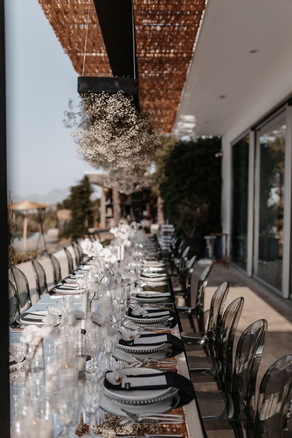 A long outdoor wedding dining table stretches along a covered terrace at Villa Amnalys in Amnatos, Crete, Greece, set with white plates edged in pearled rims, black and white napkins, and clusters of white orchids and trailing gypsophila. Clear acrylic ghost chairs line the table, and a canopy of gypsophila hangs from the woven wooden pergola above. The soft, sunlit mood is elegant and understated, with glassware catching the light.