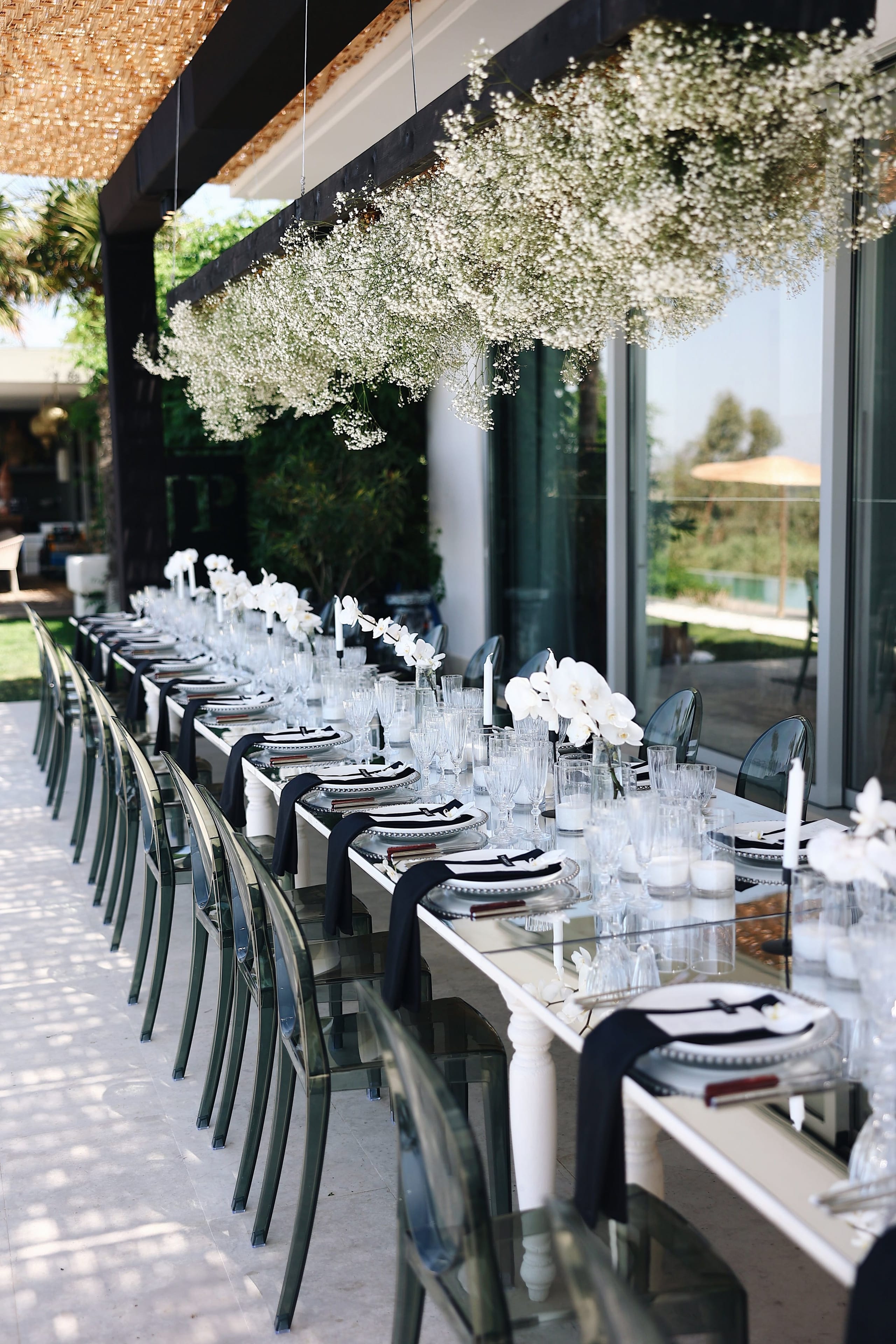 Long wedding dining table on a sunlit terrace at Villa Amnalys in Amnatos, Crete, Greece, set with white orchids, black napkins, clear glass and white candles. Above it hang two clouds of white baby's breath suspended from the dark timber beams, and smoke-grey ghost chairs line both sides of the table.
