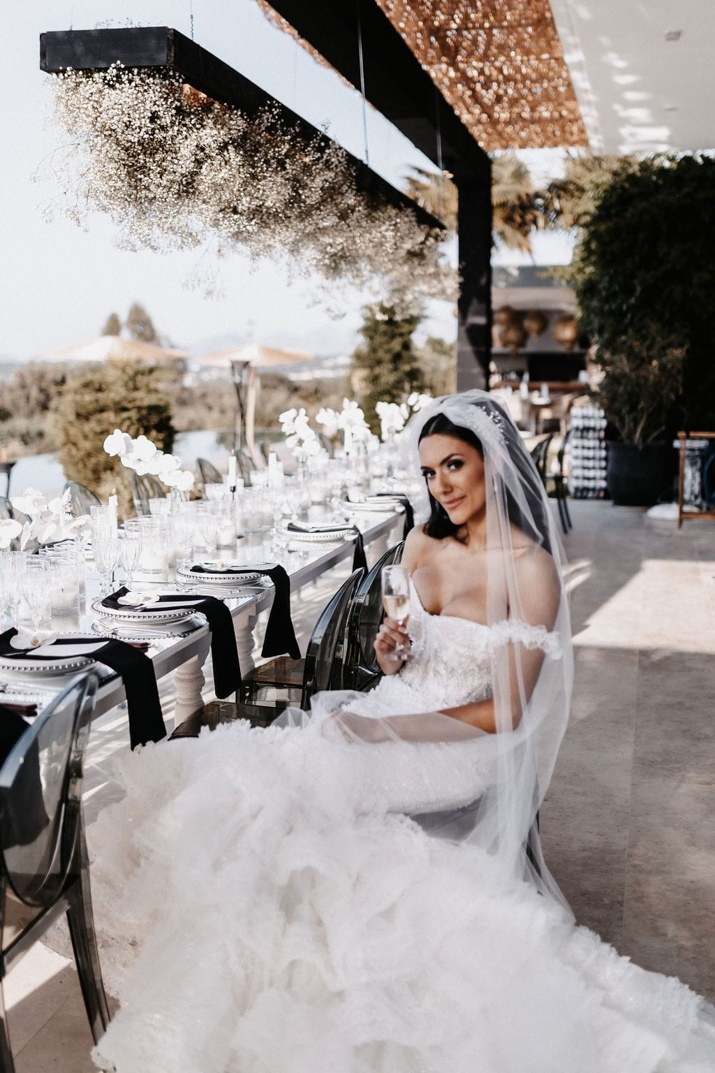 A bride in a voluminous white gown with a sheer veil sits at the head of a long terrace table at Villa Amnalys in Amnatos, Crete, Greece, holding a champagne flute and glancing back over her shoulder. The table is laid with white orchids, black runners, clear glassware and white candles, and above it hangs a cloud of dried baby's breath. Soft afternoon light falls across the stone terrace, with olive trees and a view of the sea beyond.