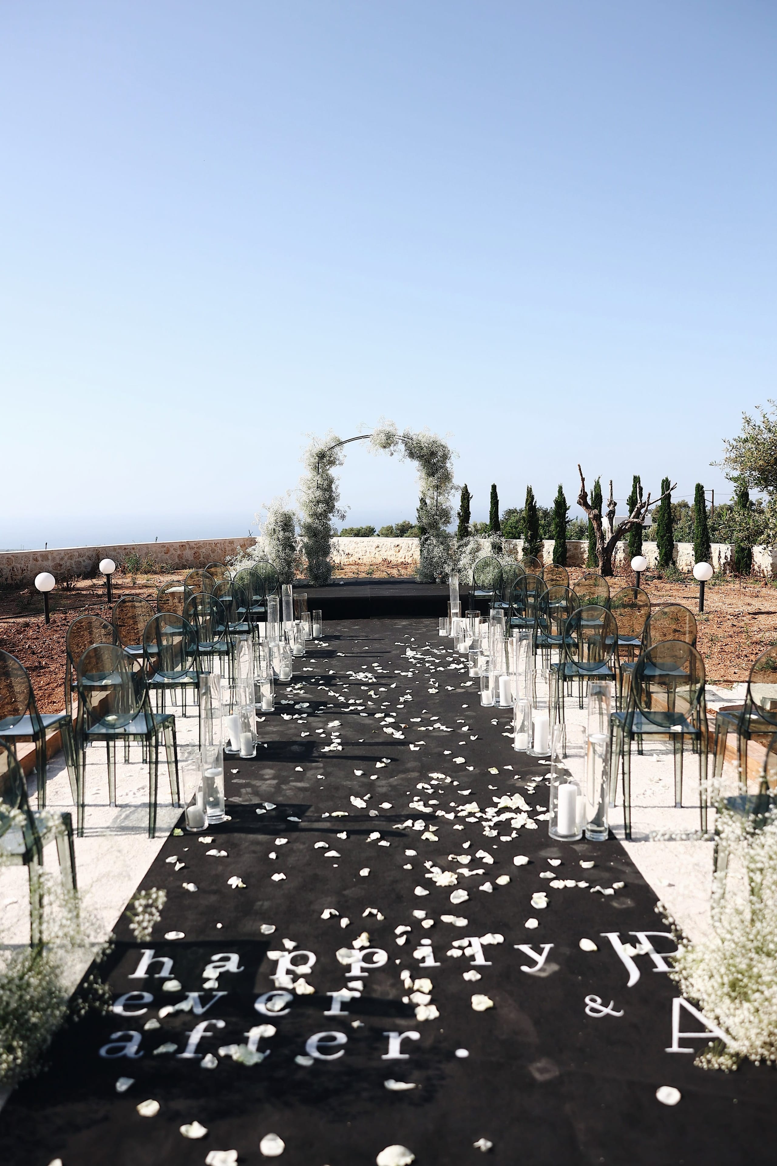 A wedding ceremony aisle at Villa Amnalys in Amnatos, Crete, Greece, running down a black runner scattered with white petals and spelling "happy ever after" in white letters. Transparent ghost chairs line both sides, with tall glass hurricane vases holding white pillar candles, and a circular arch wrapped in soft white gypsophila stands at the end under a clear blue sky, with cypress trees and an olive tree beyond the stone wall.