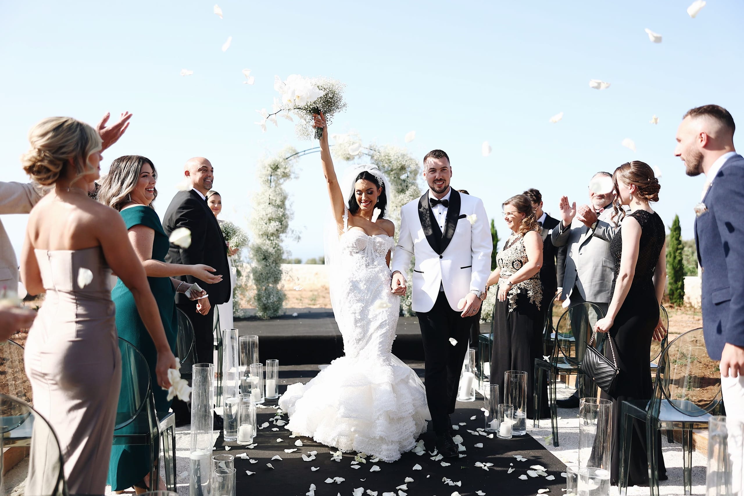 A joyful wedding recessional at Villa Amnalys in Greece, where the bride, in a fitted lace gown with a tiered train, raises a white orchid and baby's breath bouquet overhead while the groom in a white dinner jacket holds her hand. White petals drift through the air and carpet the black aisle, flanked by clear glass candle vases and guests in green and black formal wear, many smiling and applauding under a bright blue sky.