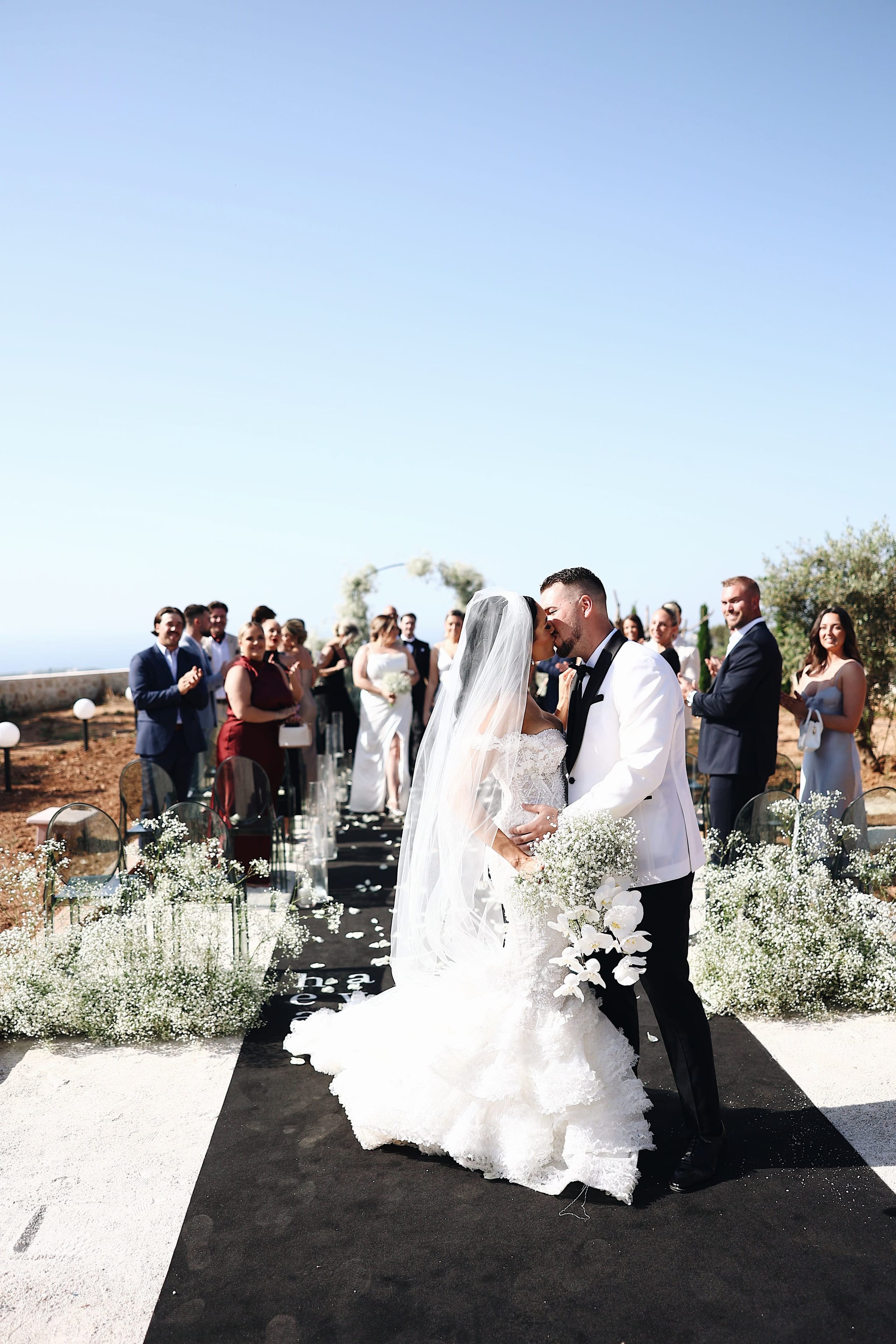 A bride in a lace gown and long veil and a groom in a white dinner jacket share a kiss at the end of a black aisle runner at Villa Amnalys in Amnatos, Crete, Greece. Clouds of baby's breath and white orchids line the aisle, and guests in pastel and deep red dresses and dark suits applaud from either side under a clear blue sky. This wedding ceremony moment is framed by olive trees.