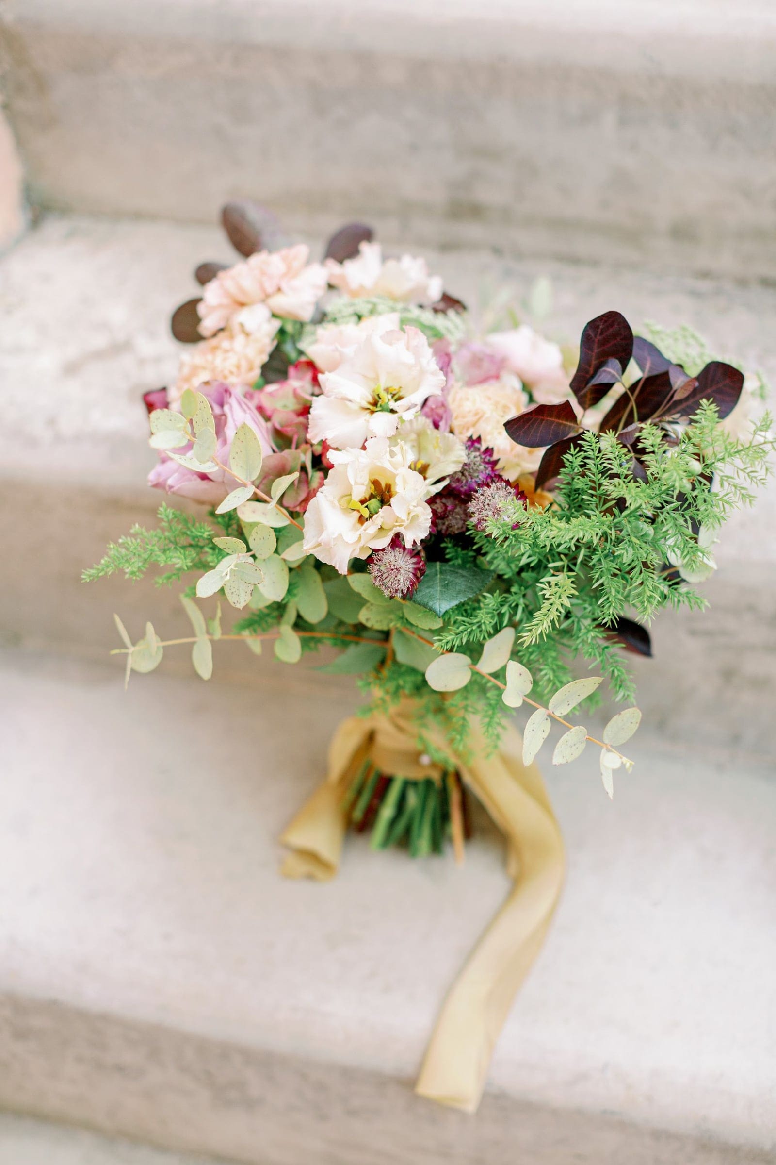 A bridal bouquet resting on pale stone steps, gathered in soft cream and blush blooms, including ivory lisianthus and peach garden roses, with purple allium heads and dark plum smoke bush leaves. Sprigs of silvery eucalyptus and feathery asparagus fern spill outward, and the stems are bound with a gold silk ribbon. This wedding flower arrangement from AgrecoFarms in Adele, Crete, Greece, has a relaxed, romantic, late-summer mood.