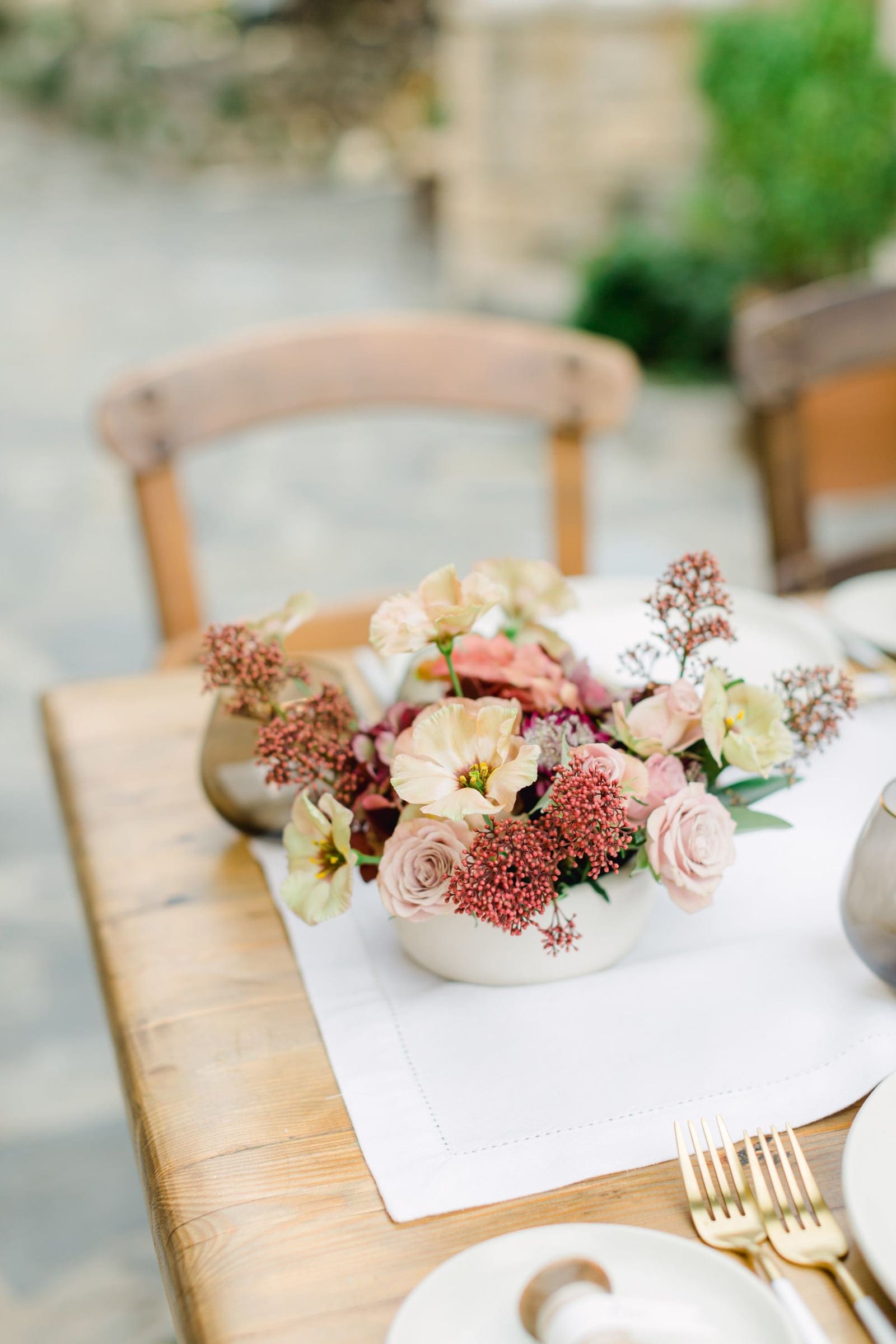 A low centrepiece in a cream ceramic bowl sits on a white linen runner atop a rustic wooden table, with blush garden roses, cream and peach poppies, and rust-red seed heads of skimmia and burgundy blooms in soft autumn tones. Gold cutlery and a pale plate sit at the edge, with wooden chairs and a blurred stone terrace behind. This is a wedding table flower arrangement at AgrecoFarms in Adele, Crete, Greece.