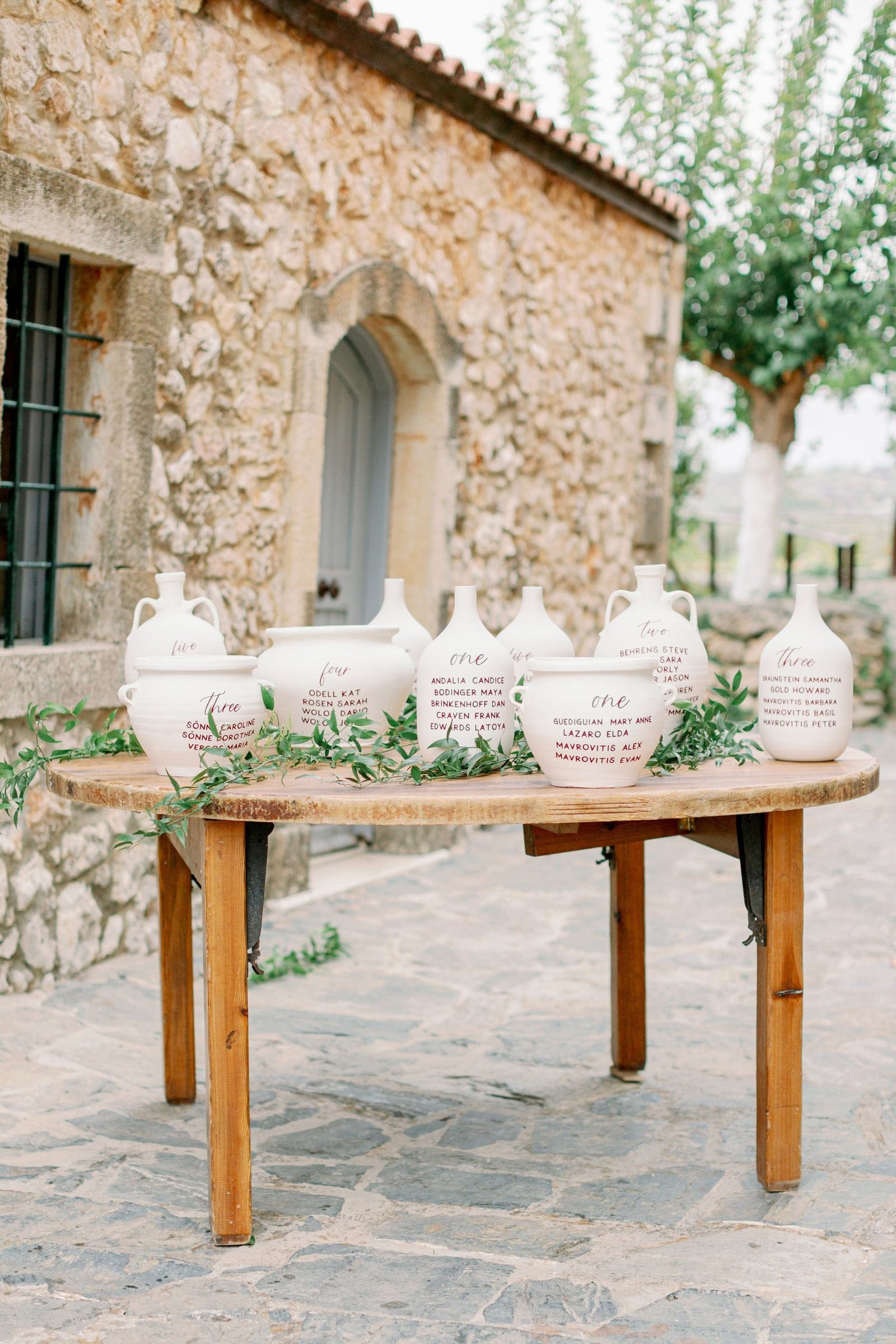 A rustic wooden table stands on a stone-paved courtyard in front of a weathered stone building, with a trailing garland of olive-like foliage draped across its top. Upon it sit a set of white ceramic jars and vases of varied shapes, each painted with a table number in elegant script and a list of guest names, forming a seating display for a wedding at AgrecoFarms in Adele, Crete, Greece.