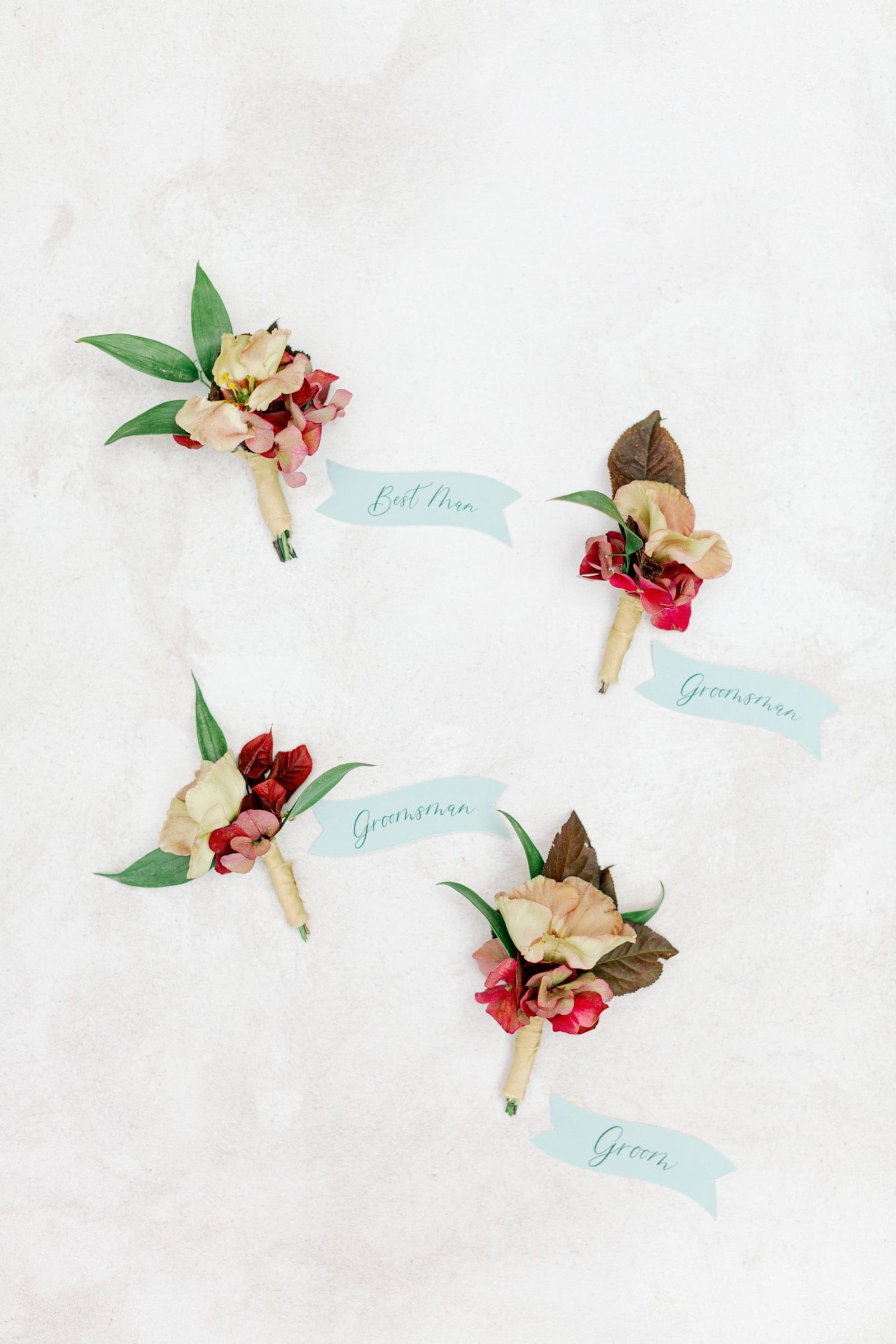 Four boutonnieres lie flat on a pale, textured stone surface, each paired with a pale blue ribbon label reading "Best Man," "Groomsman," "Groomsman," and "Groom." Each one combines a creamy, blush-edged bloom with small clusters of deep red blossoms and dark brown-green leaves, with a few slender green sprigs. These are wedding boutonnieres from Chiara and Marco's celebration at AgrecoFarms in Adele, Crete, Greece.