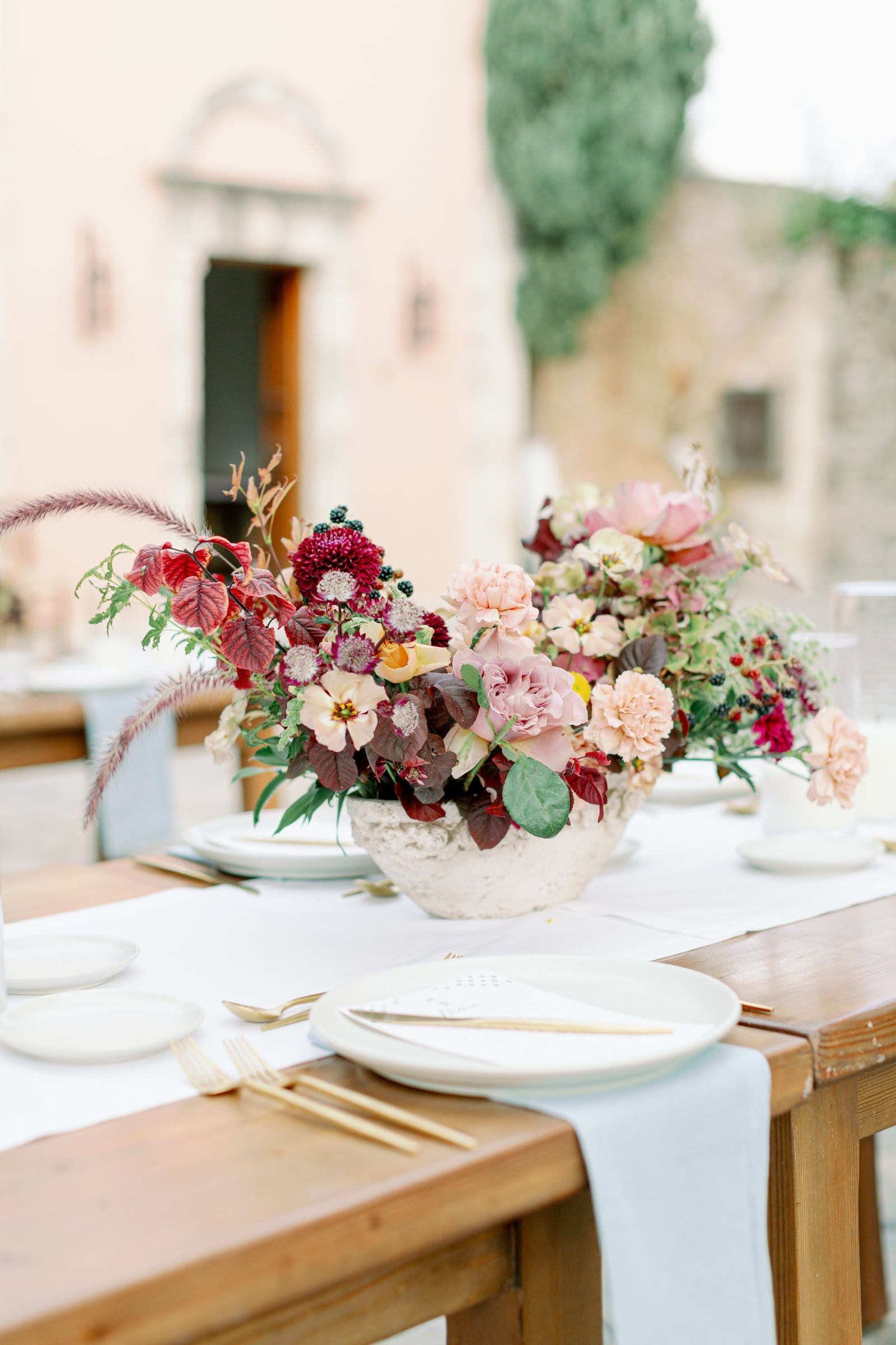 A wedding table centrepiece in a weathered cream stone bowl, filled with burgundy dahlias, blush garden roses, peach carnations, pale astrantia and dusty-pink blooms, with deep plum leaves and a feathery grass spray. It sits on a rustic wooden table dressed with a white runner, gold cutlery and white plates, in a sunlit courtyard at AgrecoFarms in Adele, Crete, Greece.