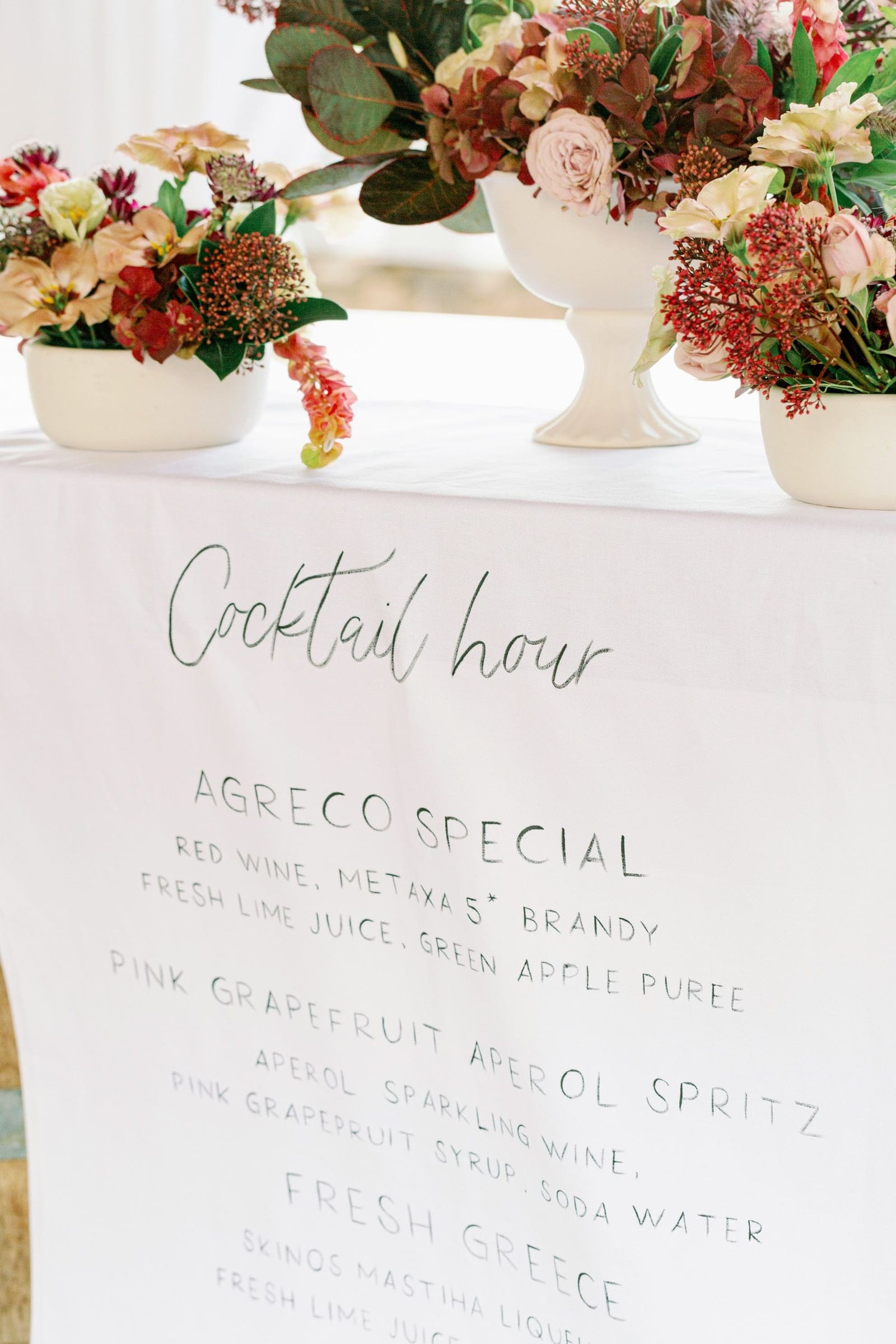 Three white footed bowls of burgundy and blush arrangements sit on a white-draped table at AgrecoFarms in Adele, Crete, Greece, for Chiara and Marco's wedding. They hold deep red hydrangea, pale pink garden roses, cream lisianthus and clusters of red berries with dark green foliage. Below them, a hand-lettered cocktail hour menu in dark green script lists drinks such as the Agreco Special and the Pink Grapefruit Aperol Spritz.