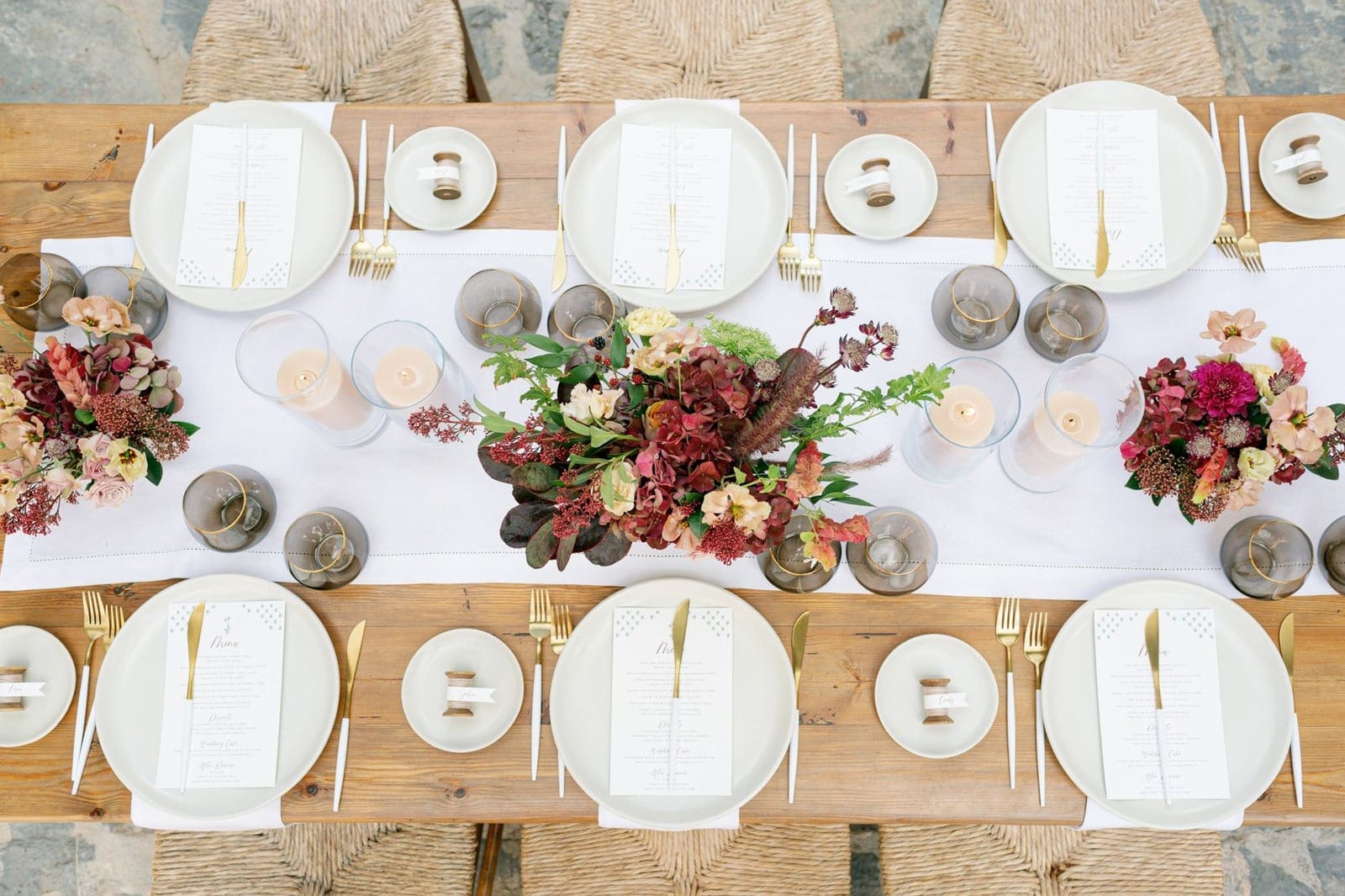 A long rustic wooden wedding table at AgrecoFarms in Adele, Crete, Greece, viewed from above, dressed with a white linen runner. A dense centrepiece of deep burgundy hydrangeas, pale peach roses and dark seed heads sits in the middle, flanked by smaller arrangements of blush and mauve blooms, with pale candles in glass hurricanes and smoky grey glasses rimmed in gold. Each place setting has a white plate, gold cutlery and a printed menu card, and the woven rush chairs at the ends suggest a relaxed, earthy mood.