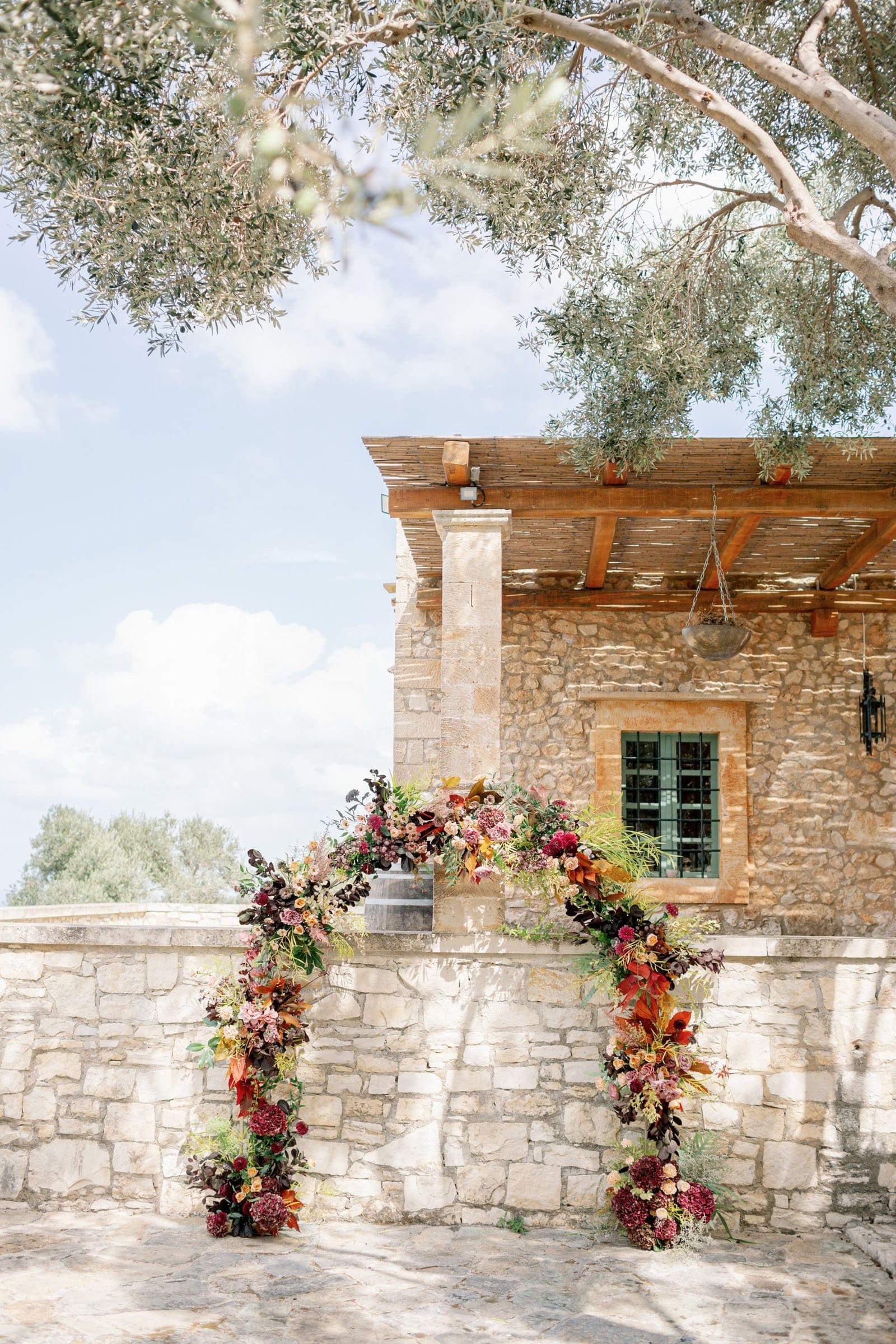 A wedding arch of deep burgundy and rust-toned blooms, with dark hydrangeas, pink roses and trailing autumn foliage, curves over a low stone wall at AgrecoFarms in Adele, Crete, Greece. Behind it stands a warm honey-coloured stone farmhouse with a wooden pergola and a green-framed window, while olive branches overhead filter soft, pale sunlight onto the pale flagstones below.