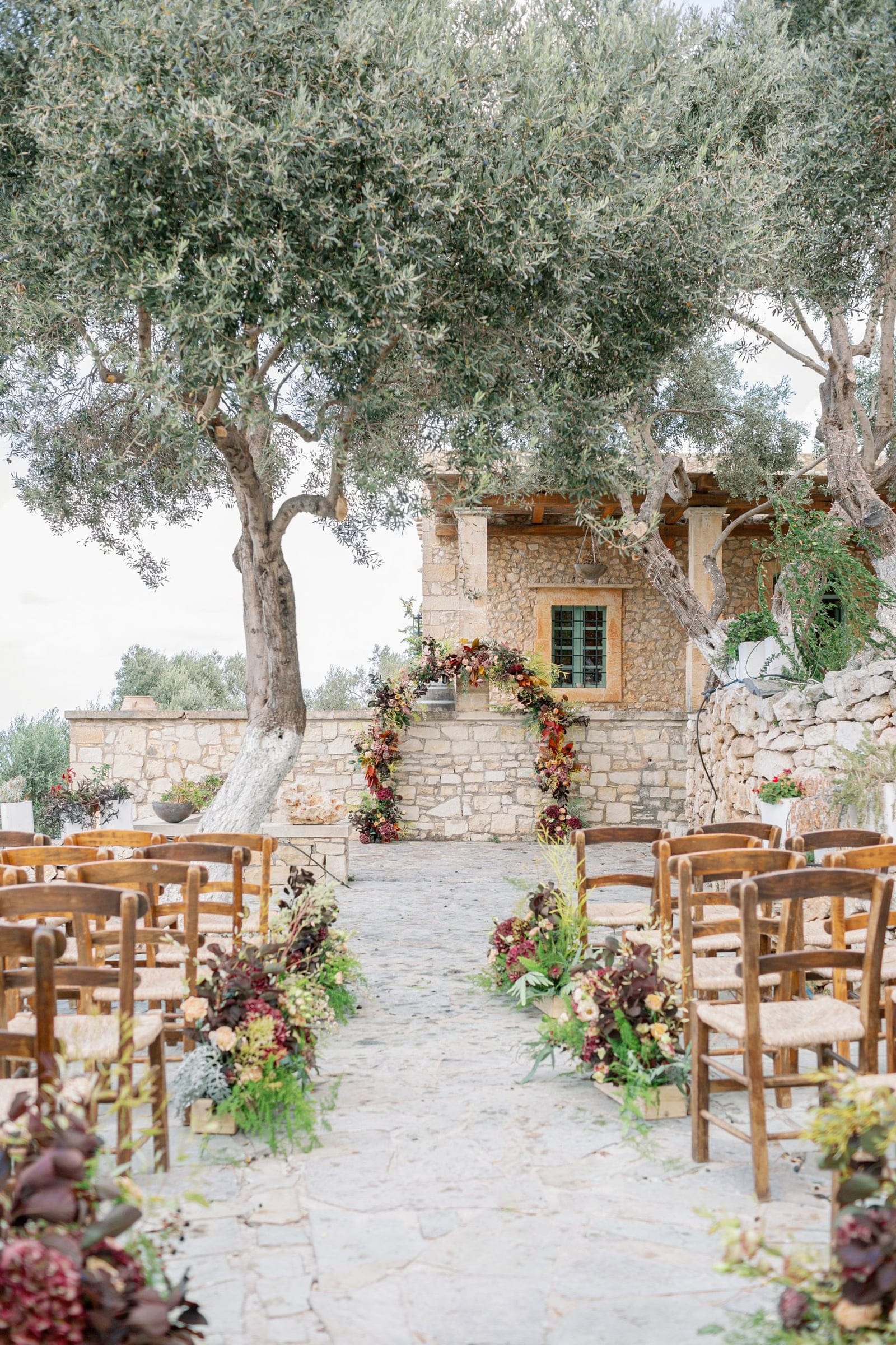 A wedding ceremony aisle in Greece, lined with rows of rustic wooden chairs on a flagstone path under large olive trees. At the far end, a curved arch of deep burgundy and rust-toned blooms and foliage frames a stone wall at AgrecoFarms in Adele, Crete, where wedding flowers sit in low wooden boxes along both sides of the aisle. The soft, overcast light and muted tones give the scene a calm, autumnal mood.