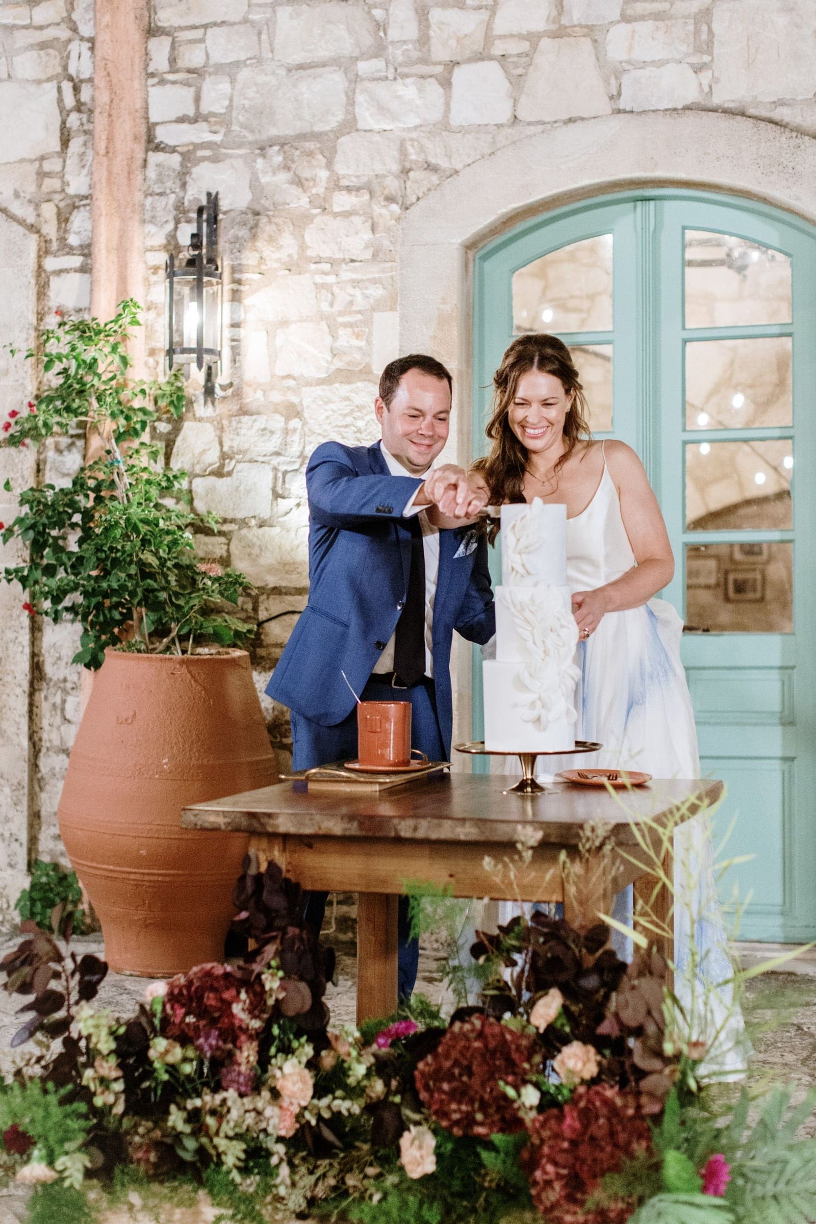 A bride in a white satin slip dress and a groom in a navy suit cut a three-tier white cake together, both smiling, at a wooden table in a stone-walled courtyard at AgrecoFarms in Adele, Crete, Greece. Beside them, a large terracotta pot holds a climbing bougainvillea, and the table is framed by a low arrangement of deep burgundy hydrangeas, peach roses and dark foliage. Behind them, a pale teal arched door and a wall lamp add a warm, rustic glow.