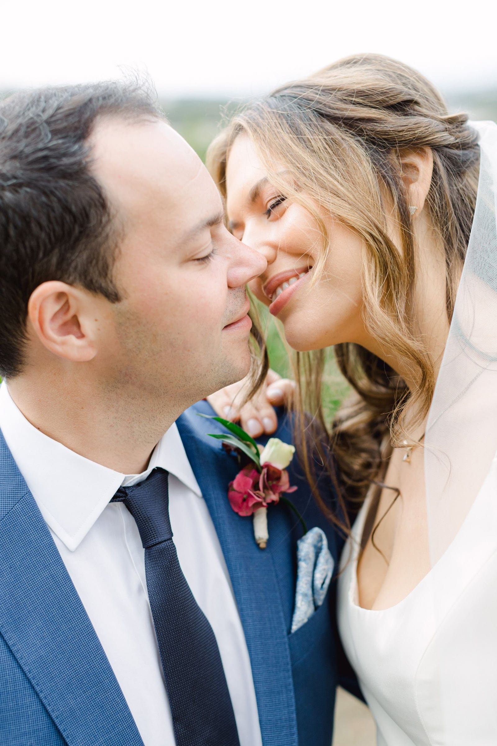 Close-up of a bride with soft wavy hair and a sheer veil, smiling with her eyes half-closed as her nose nearly touches that of a groom in a navy suit and white shirt, his eyes closed. A small boutonniere of white and burgundy blooms with green foliage is pinned to his jacket, and the blurred green landscape behind them suggests an outdoor wedding at Agreco farm in Greece.