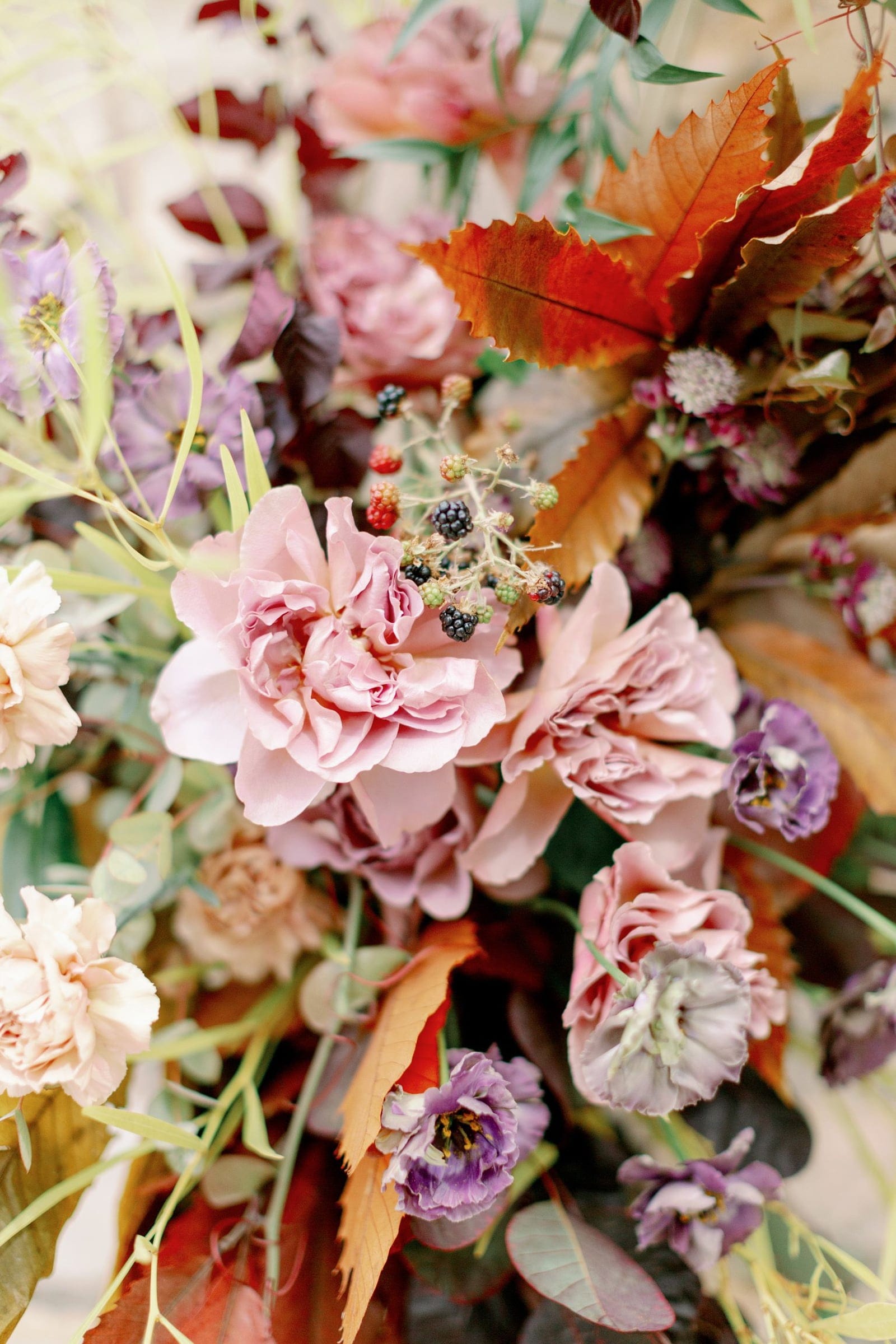 Close-up of a wedding arrangement at AgrecoFarms in Adele, Crete, Greece, built around dusky pink garden roses with ruffled petals. Burnt-orange and rust chestnut-like leaves, deep plum foliage, purple lisianthus, small blackberries and pale astrantia weave through the bouquet, with soft cream carnations and silvery eucalyptus at the edges. The warm autumnal palette and gently blurred background give it a rich, romantic feel.