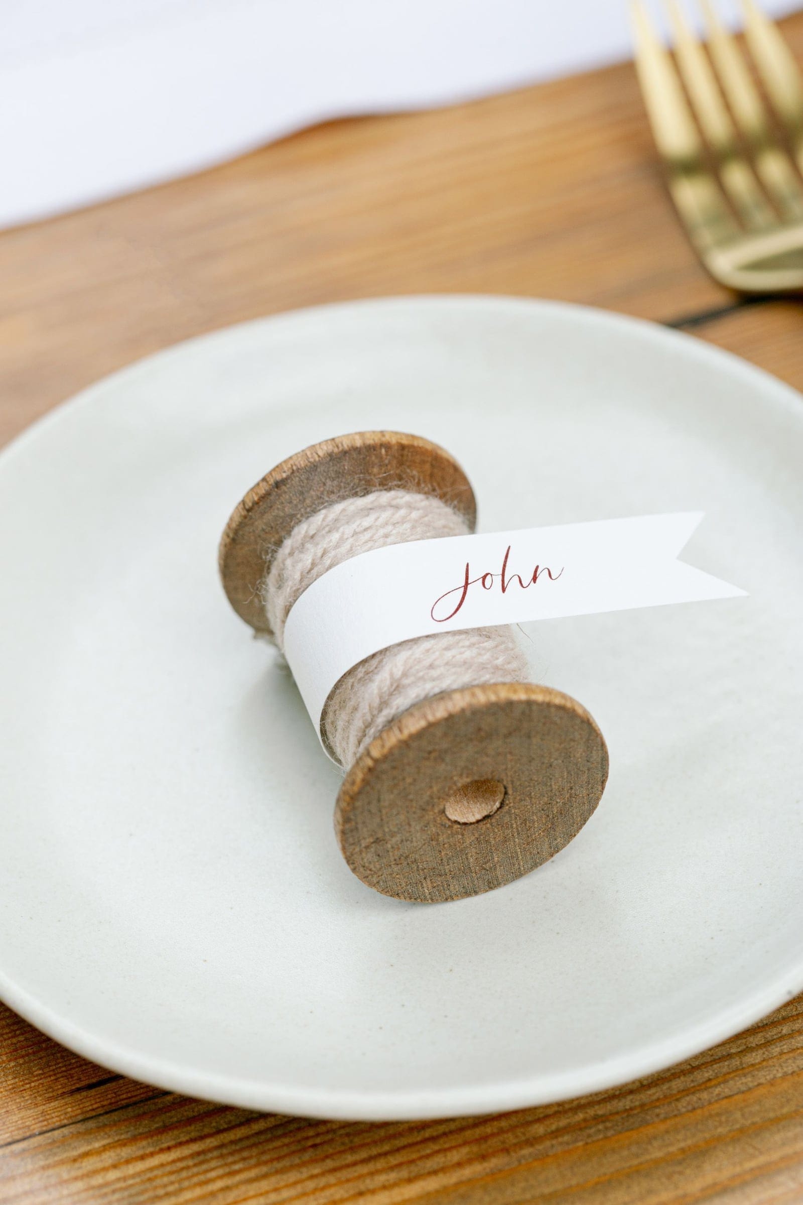A rustic wooden spool wrapped in soft beige twine sits on a pale grey-green plate, with a white paper flag tied around it bearing "John" in red script calligraphy. Gold cutlery rests on the wooden table in the upper corner. These are wedding place settings from Chiara and Marco's celebration at AgrecoFarms in Adele, Crete, Greece.