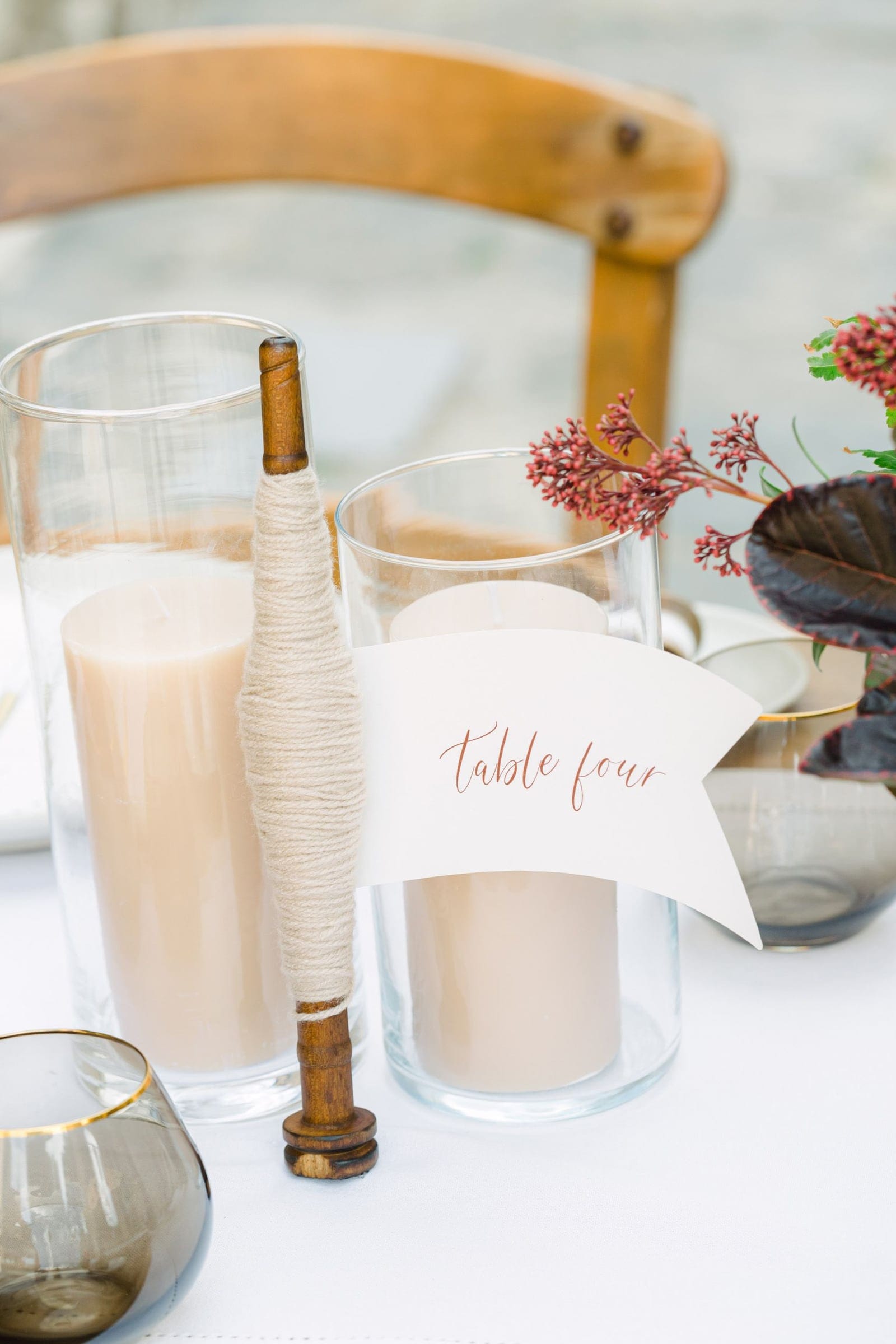 A wedding table setting at AgrecoFarms in Adele, Crete, Greece, dressed in warm neutrals. Two clear glass cylinders hold pale beige pillar candles, and a wooden stick wrapped in natural twine stands between them. A white flag-shaped card in delicate rust-coloured script reads "table four." At the right, a dark bronze-leaved stem with small rust-red berry clusters rises from a smoked glass with a gold rim, and a wooden chair sits softly blurred behind the table.
