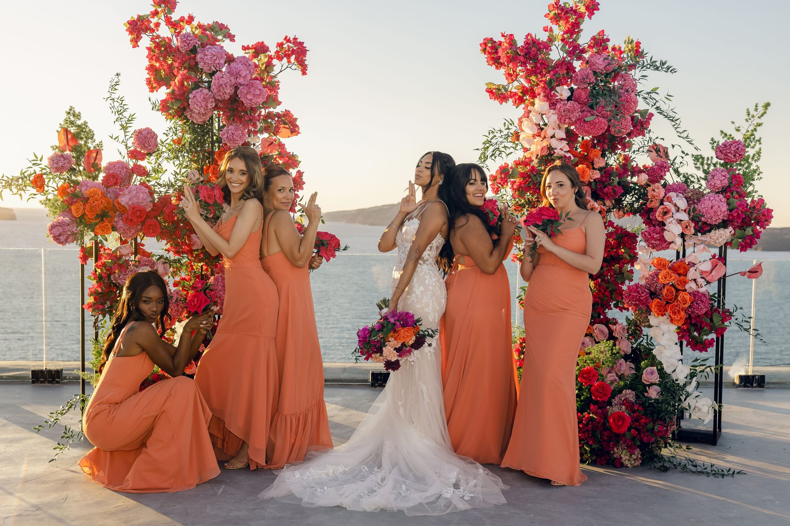 A bride in a fitted white lace gown stands in the centre of five bridesmaids in coral-orange dresses, all posing in front of tall floral installations of hot pink hydrangeas, red bougainvillea and orange roses on a clifftop terrace at Cavo Ventus Luxury Villa in Santorini, Greece. Her bouquet is a mix of purple, orange and blush blooms, and the soft evening light over the sea makes the whole wedding scene glow.
