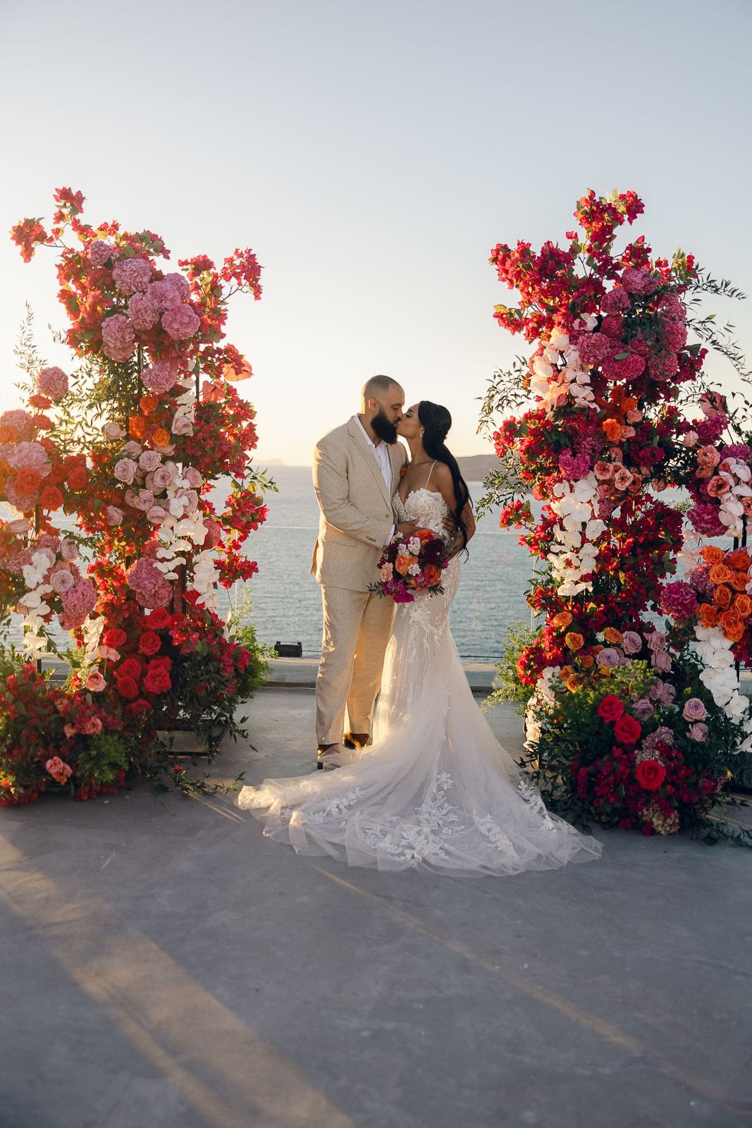 Two tall floral pillars frame a bride and groom kissing at Cavo Ventus Luxury Villa in Santorini, Greece, with the sea and soft sunset light behind them. The arches are packed with bougainvillea, pink and red roses, peonies, white orchids and orange blooms, while the bride holds a matching bouquet of coral and deep red flowers. She wears a fitted lace gown with a long train, and he wears a light beige suit.
