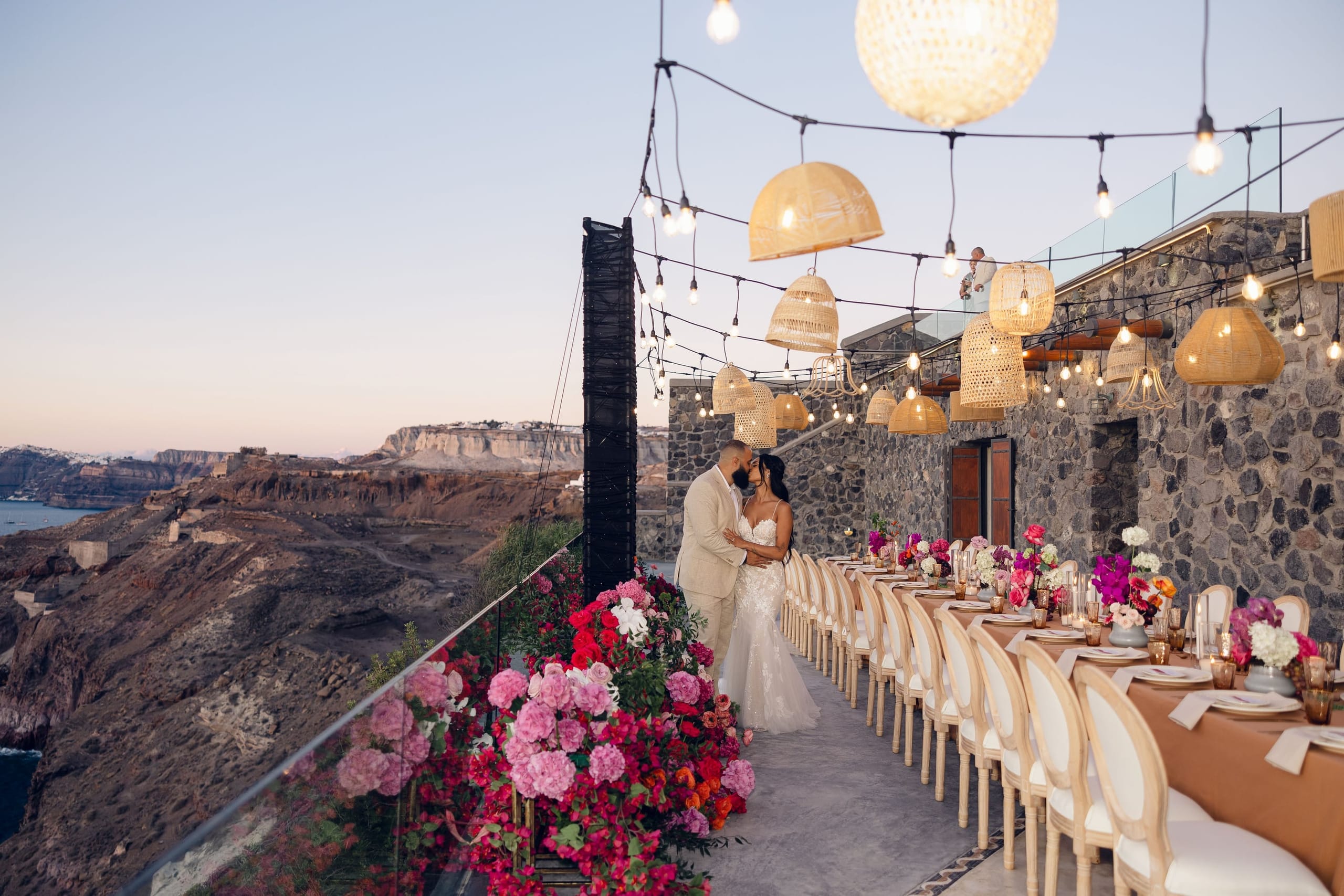 A bride in a fitted lace gown and a groom in a cream linen suit share a tender kiss at dusk on a stone terrace edged by a glass railing, with a long wedding table running beside them. Along the railing, cascades of pink peonies, fuchsia and magenta bougainvillea, and red blooms spill over lush greenery, while wicker pendant lamps and strings of bulb lights glow overhead above the caldera cliffs. This is a wedding setting at Cavo Ventus Luxury Villa in Santorini, Greece.