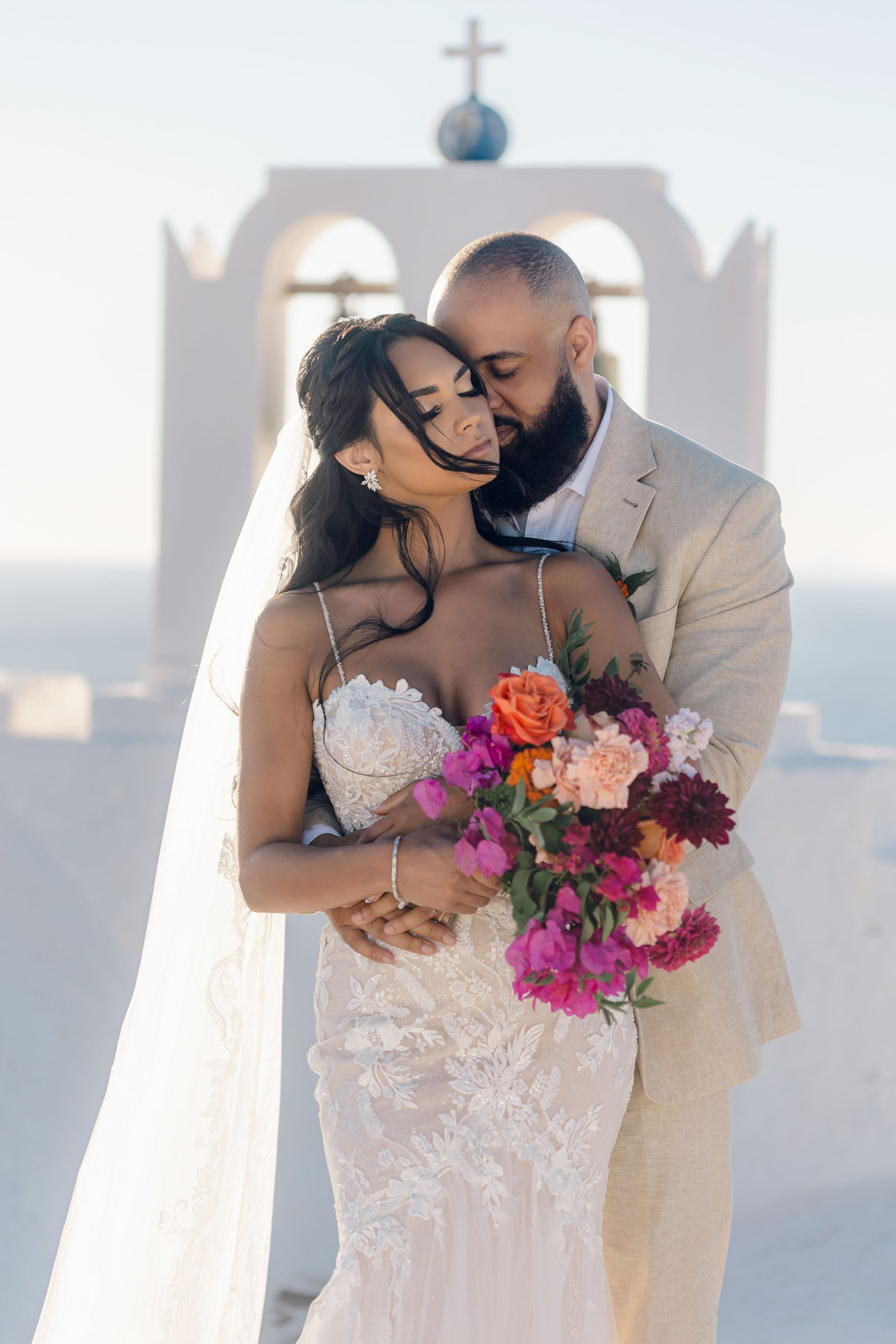 A bride with dark hair in a loose side braid and a lace-bodice gown with thin straps stands with her eyes closed while a bearded groom in a light linen suit leans in and rests his forehead against hers. She holds a bright bouquet of orange roses, pink carnations, magenta bougainvillea and deep burgundy dahlias with green foliage. Behind them, white arched bell towers glow in soft sunlight over a pale sea, marking this wedding portrait at Cavo Ventus in Greece.