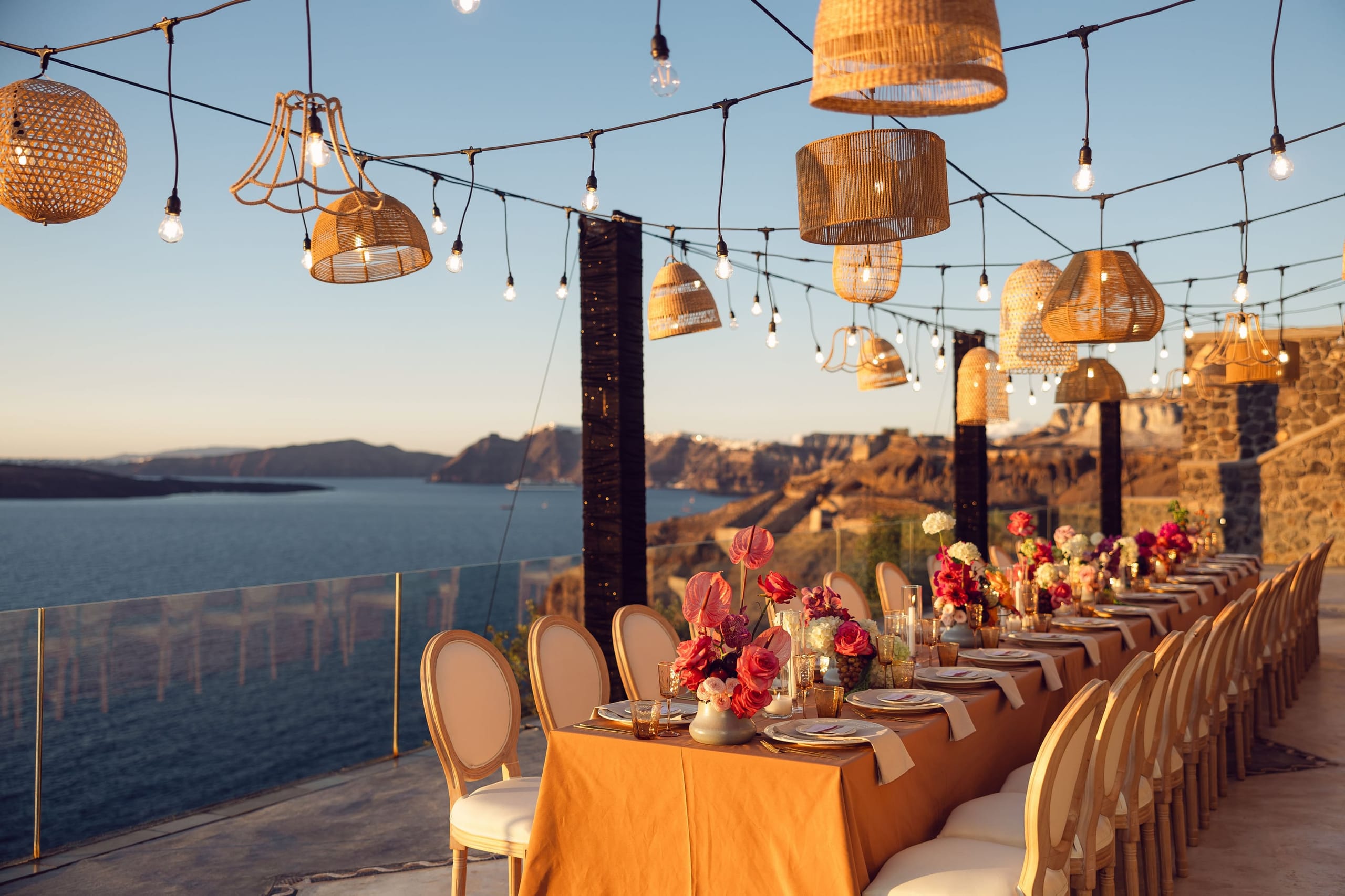 A long wedding dinner table draped in burnt-orange linen runs along a clifftop terrace at Cavo Ventus Luxury Villa in Santorini, Greece, at golden hour. Its centrepieces mix pink anthuriums, deep red roses and white blooms, and gold-rimmed glasses and cream chairs line both sides. Woven rattan pendant lampshades and strings of bulb lights hang overhead above the table, and beyond the glass railing the caldera sea and rugged cliffs stretch into the warm evening haze.